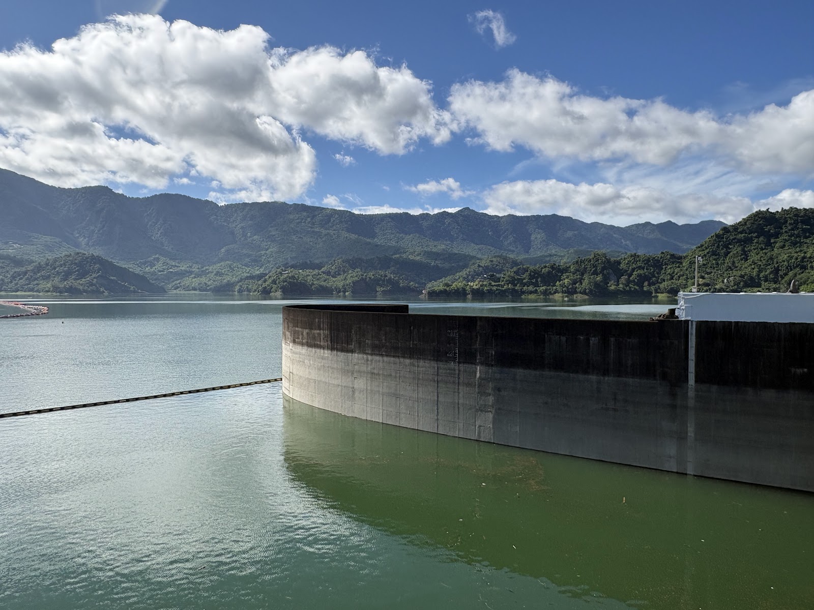 曾文水庫半圓形溢洪道消力池近景，背景碧綠湖水和青翠山巒層疊，天空藍白
