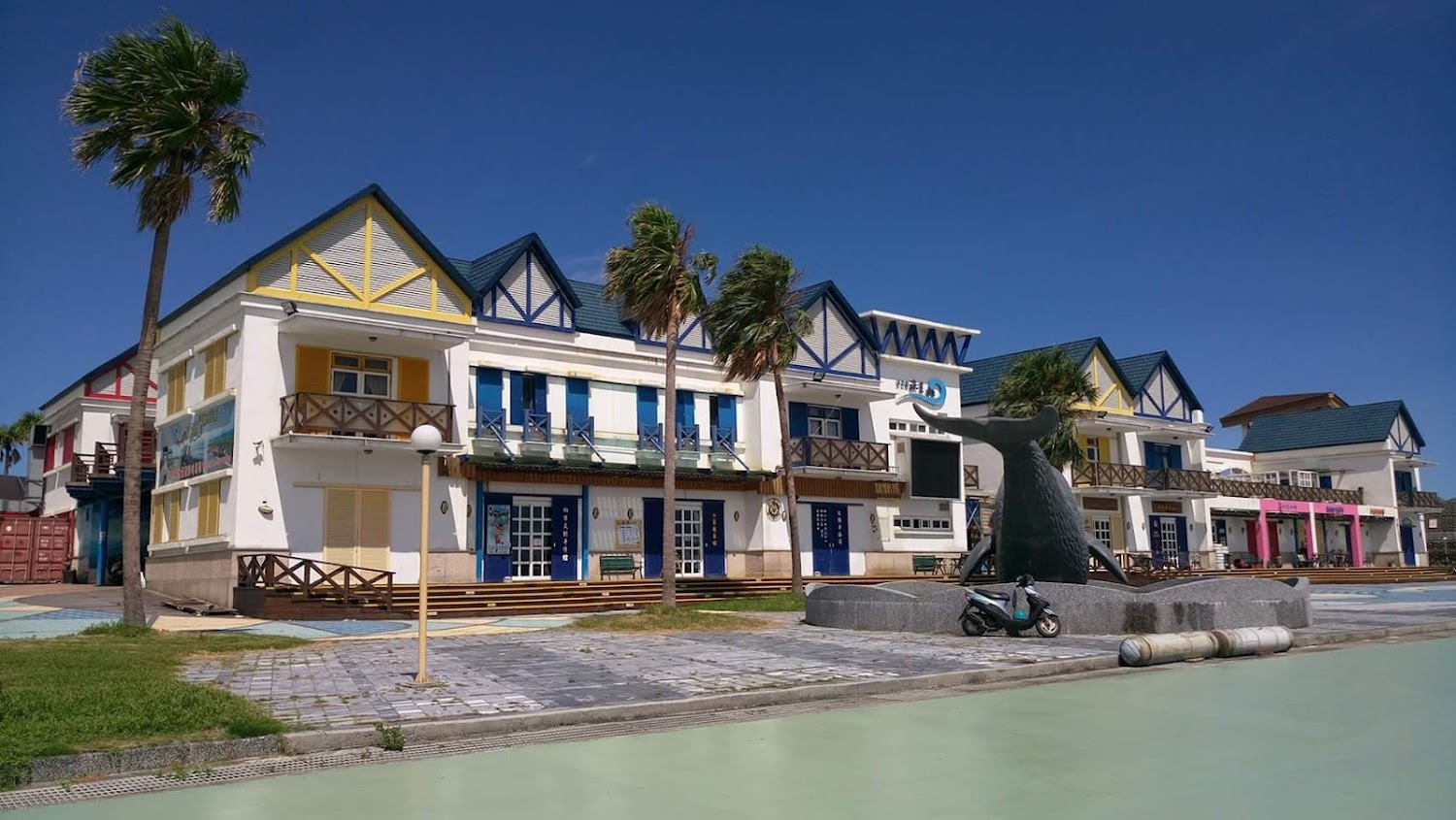 這張圖片展示了一排色彩鮮豔的歐風建築,前方有棕櫚樹和一個鯨魚尾巴造型的雕塑,還有一台機車停放,天氣晴朗,沒有明顯浮水印。