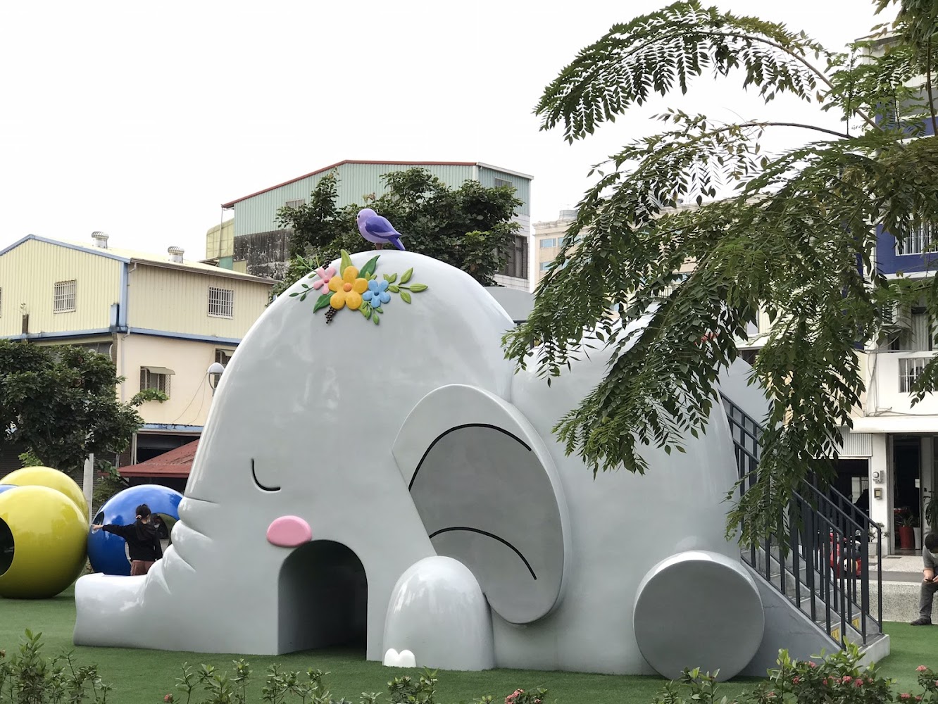 這是一座設計成大象造型的兒童遊樂設施,大象頭上裝飾有花朵和一隻鳥,旁邊有樓梯可供攀爬。