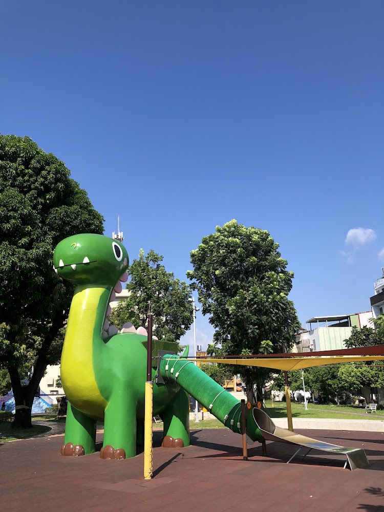 這張圖片主要是一個綠色恐龍造型的溜滑梯遊樂設施,周圍有樹木和建築物,天空晴朗,畫面色彩鮮明。