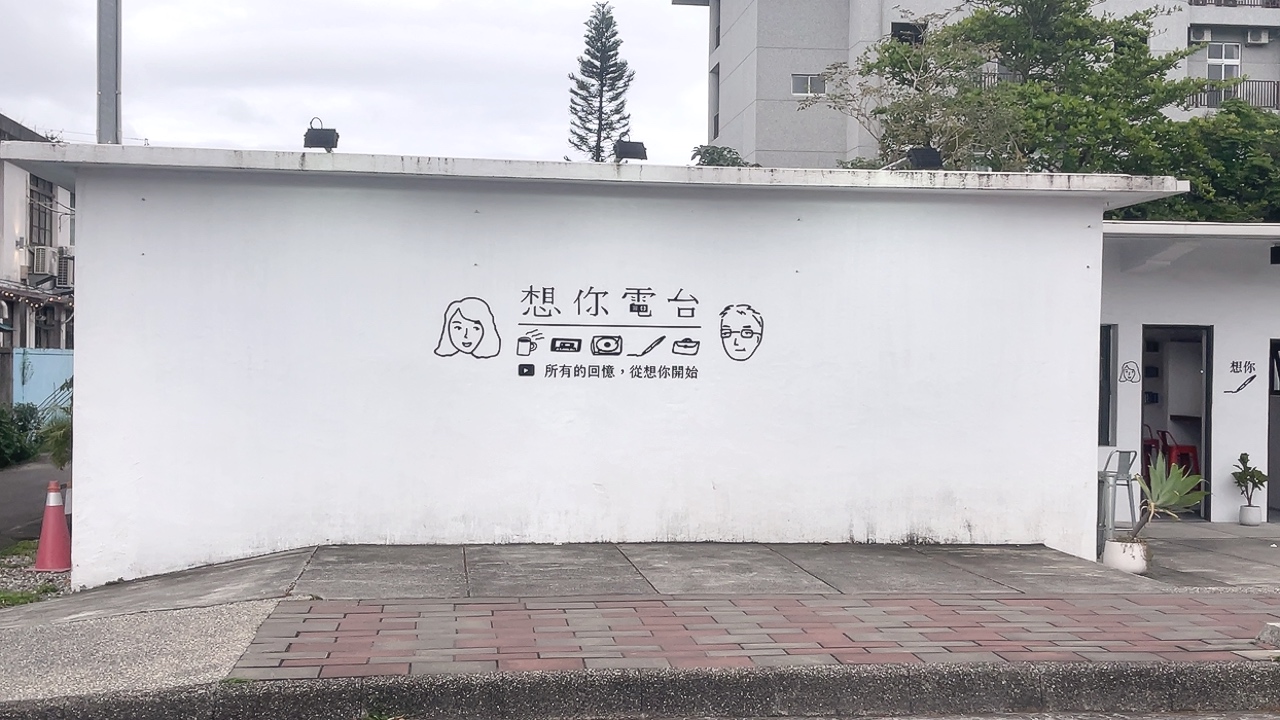 這張圖片顯示一棟白色建築,牆上有『想你電台』標誌與插畫,旁邊還有中文字與影音、飲品等圖案。現場氛圍簡約溫馨,沒有明顯浮水印。