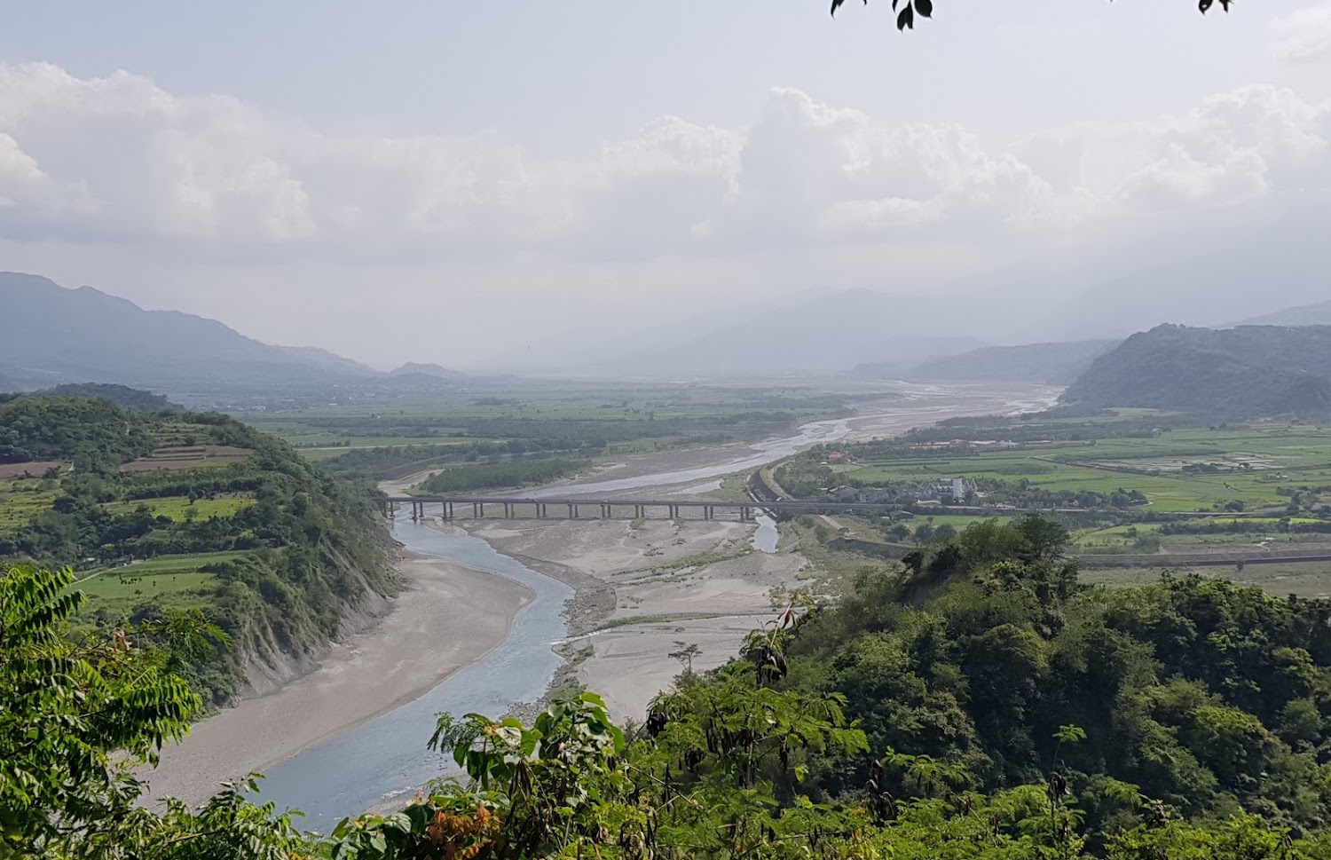 河流穿越綠意山谷，河上有橋樑和遠方山脈