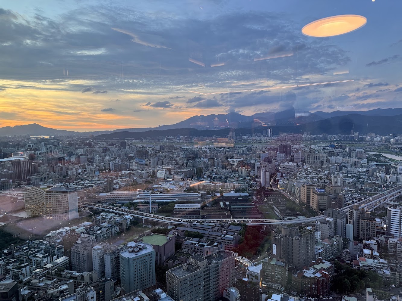 城市高空景觀，可以看到許多建築和道路，遠方有山脈和夕陽天空