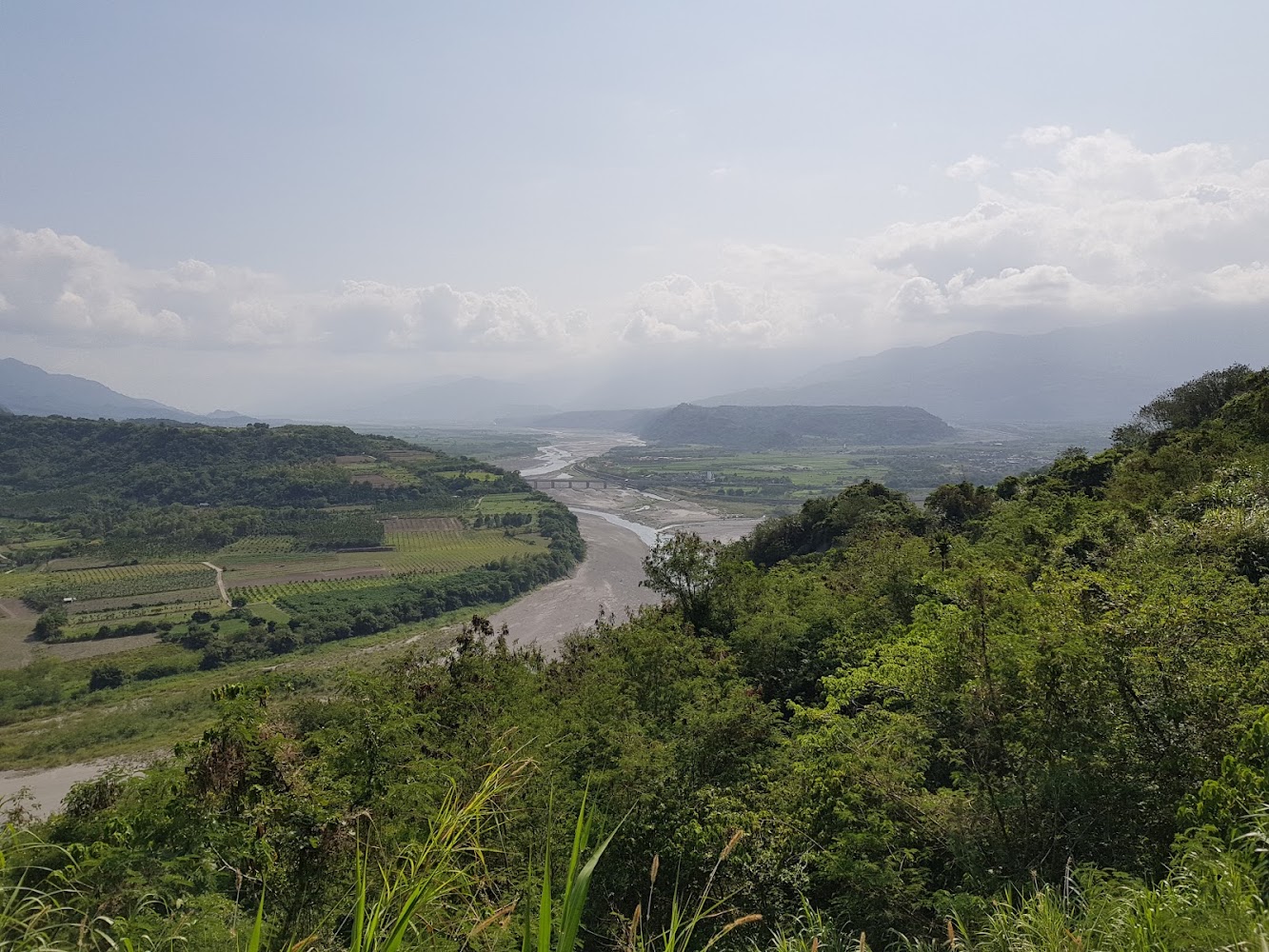 河流蜿蜒穿過青翠山谷與田野，遠方有橋樑和山脈