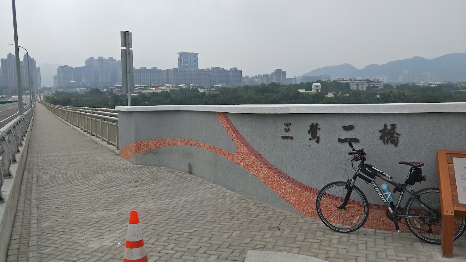 寫有三峽二橋名稱的橋梁旁停著自行車，遠方可見都市高樓與山景。