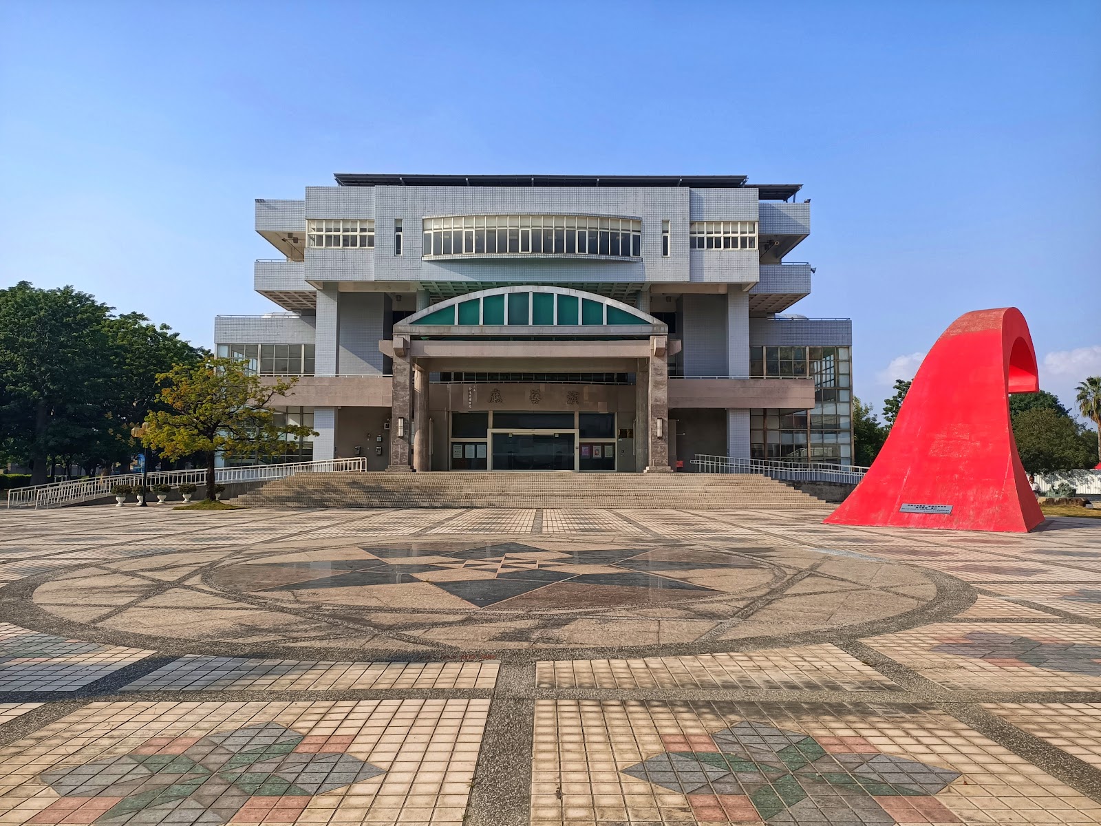 岡山文化中心正面外觀，紅色弧形裝置藝術與廣場地磚圖案，高雄岡山人文景點