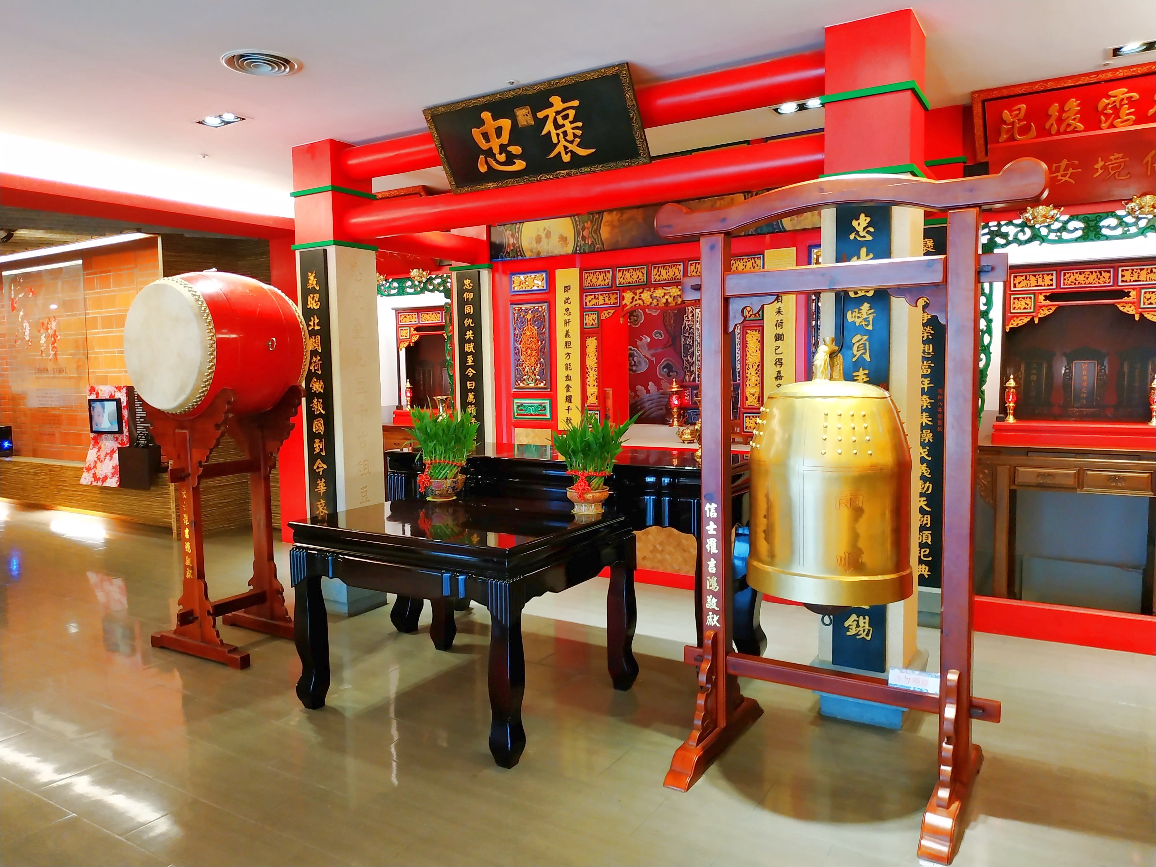 這張圖片主要展示了中式廟宇風格的室內空間，中央有一面大鼓和一口金色大鈴，周圍有紅色柱子與展示牌匾，整體裝飾充滿傳統文化氛圍。圖片無明顯浮水印。