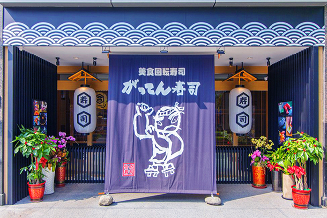 這是一家日式壽司店的店面，門口懸掛著大布簾，上面有日本字樣和武士圖案，兩側有大型燈籠與盆栽裝飾，充滿日本風情，現場沒有浮水印。