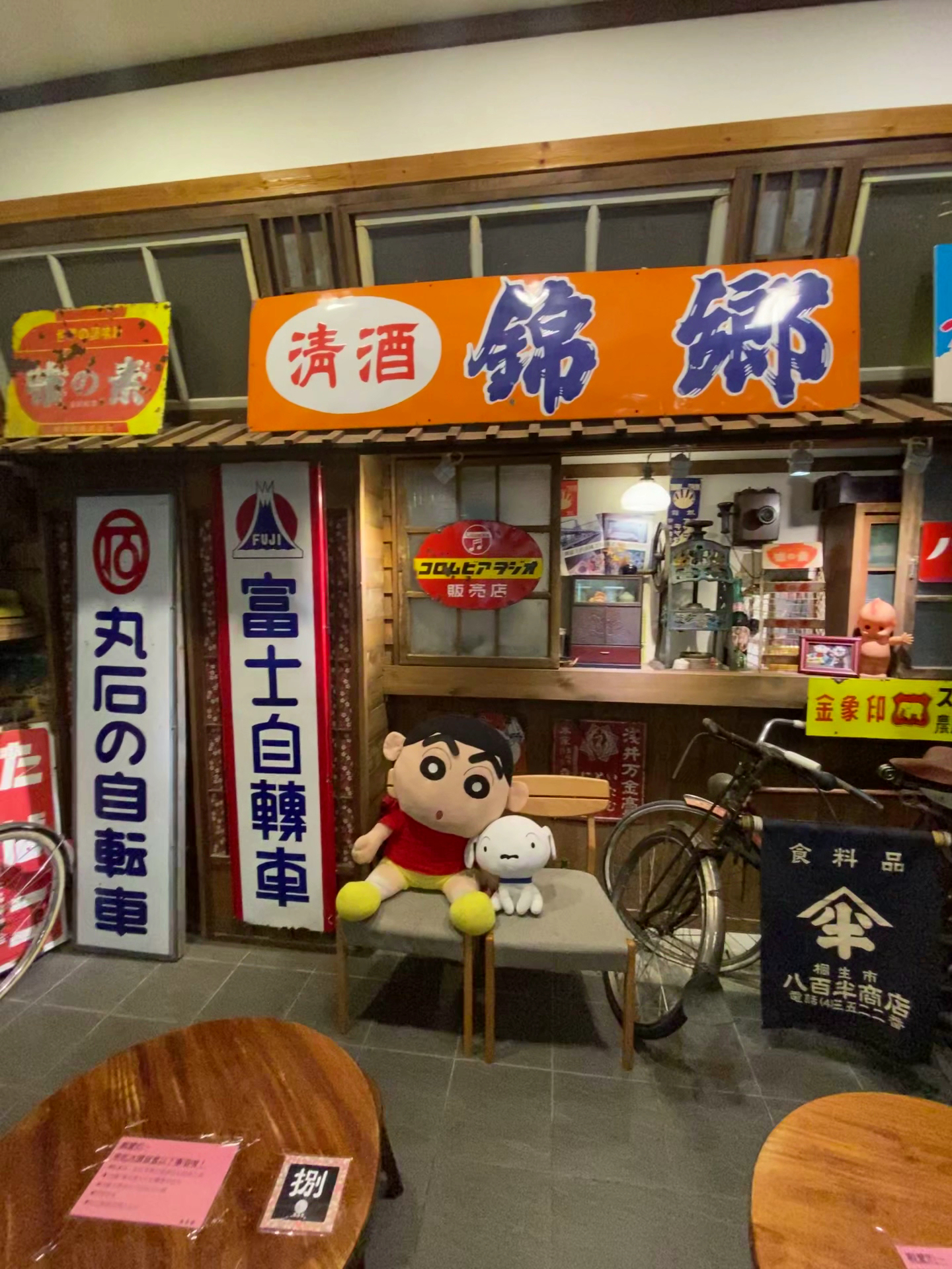這張圖片展示一個具有昭和時代風格的日式復古店面,門口有蠟筆小新和小白的玩偶、懷舊廣告招牌、自行車,以及一些舊時物品,營造出懷舊氣氛。圖中無明顯浮水印。