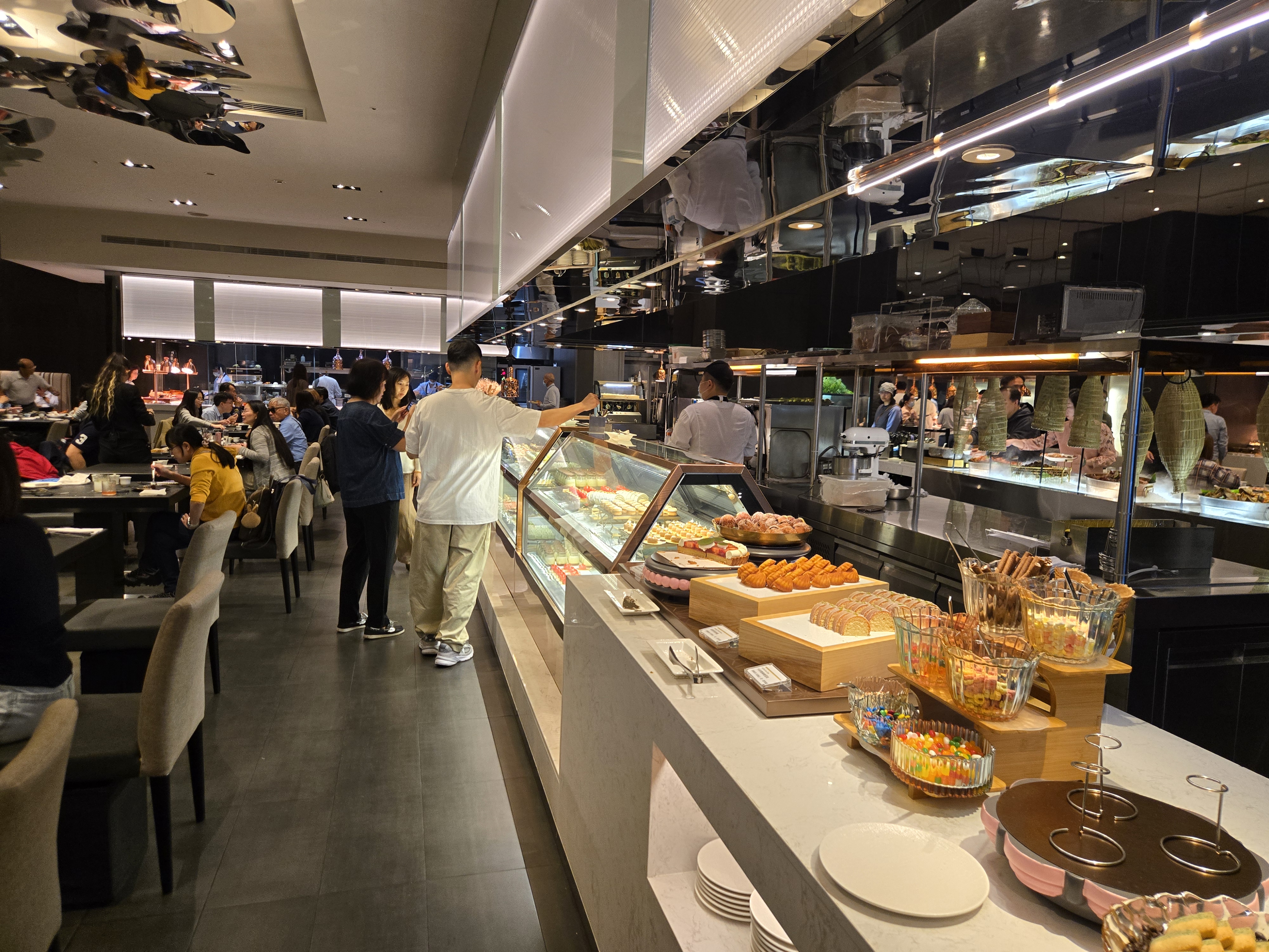 這張圖片主要展示了一家自助餐廳，前方有各式甜點和糖果，後方有顧客正在取餐，環境明亮且熱鬧。未見明顯浮水印。
