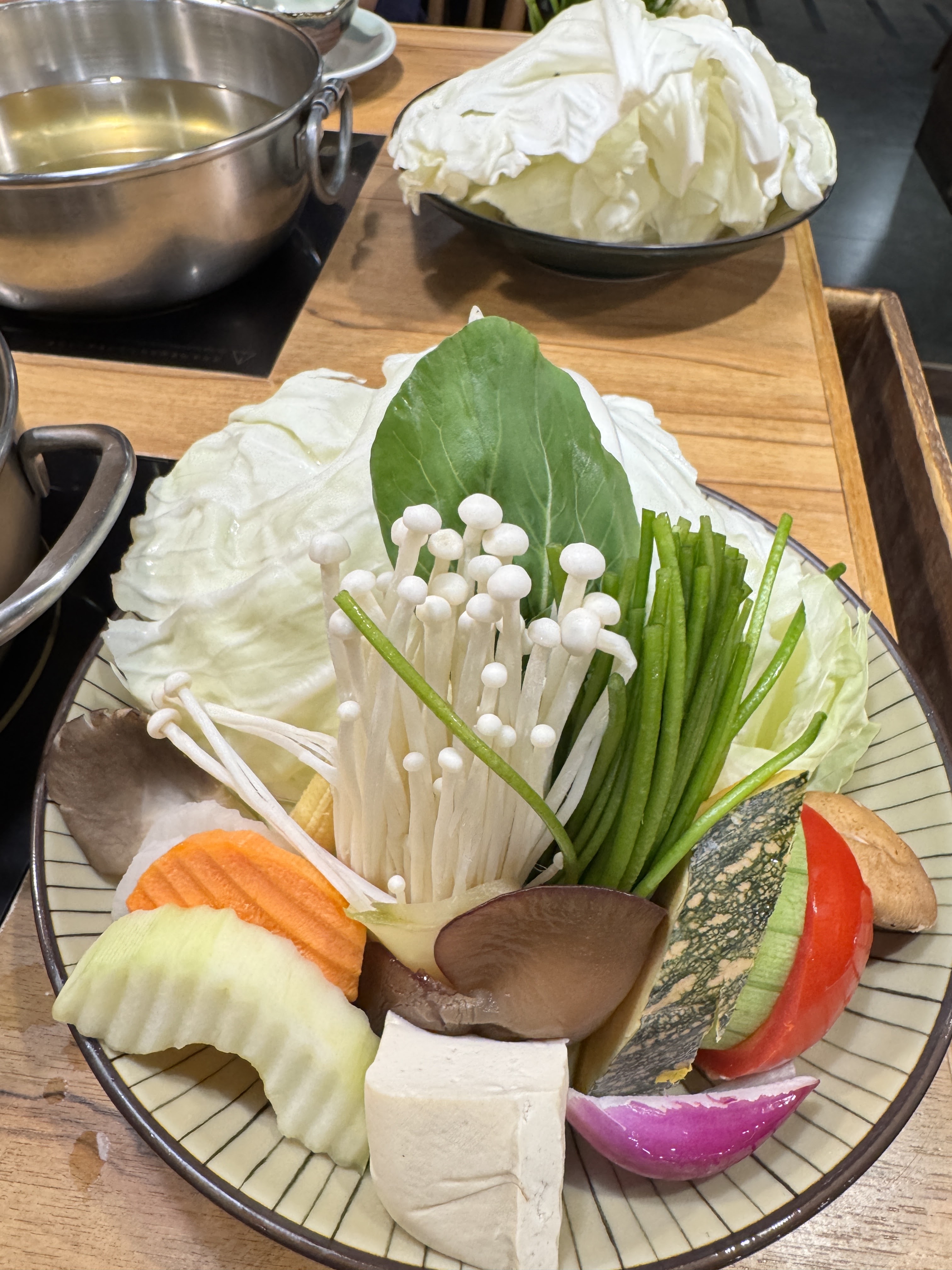 這張圖片展示一盤火鍋蔬菜拼盤,包含高麗菜、金針菇、香菇、南瓜、番茄、豆腐、青菜、紅蘿蔔等,後方還有湯鍋和更多高麗菜。圖片無浮水印。