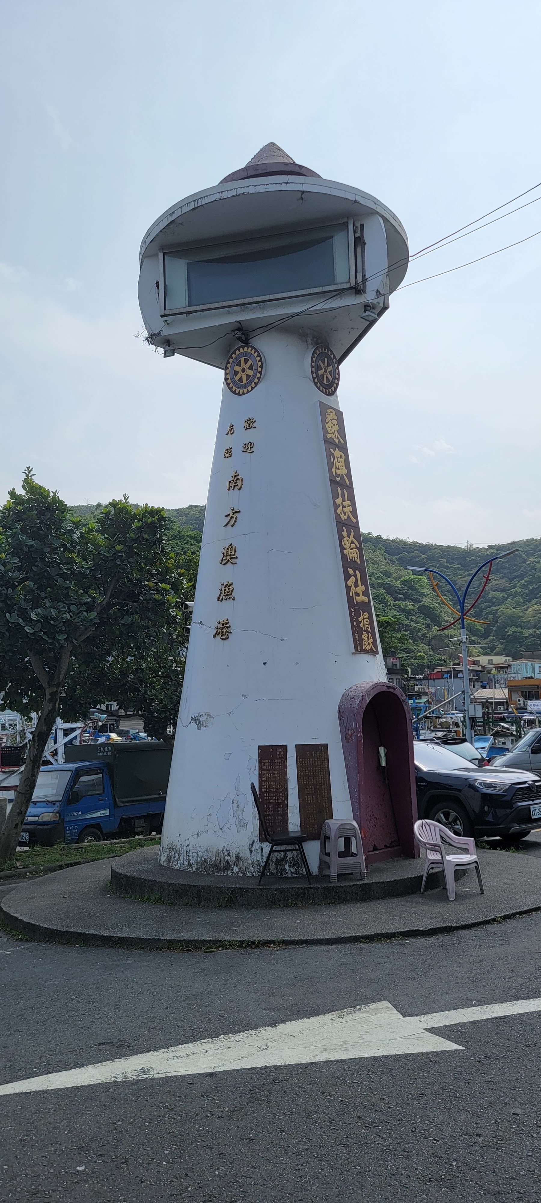 這張圖片的主要物件是一座白色燈塔造型的建築，表面有金色中文字，寫著『歡迎光臨南方澳漁港』等。周圍擺放有幾張椅子，背景可以看到停放的車輛及山景。圖片中無明顯浮水印。
