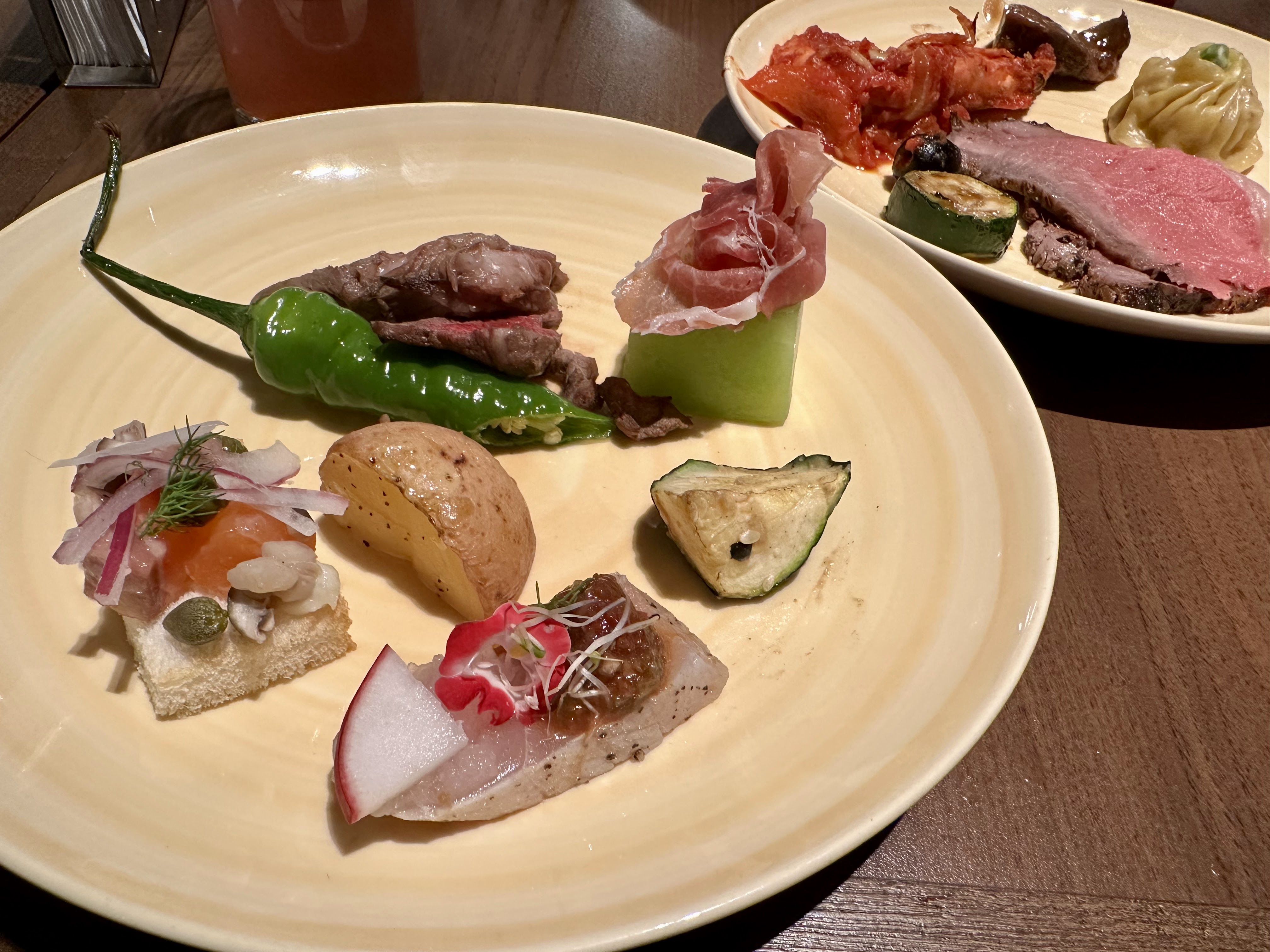 主盤有烤牛肉、瓜片、生火腿哈密瓜、馬鈴薯、青辣椒、煙燻鮭魚小吃及裝飾用蘿蔔、小菜等。後方還有泡菜、蒸餃與肉片。圖片沒有明顯浮水印。