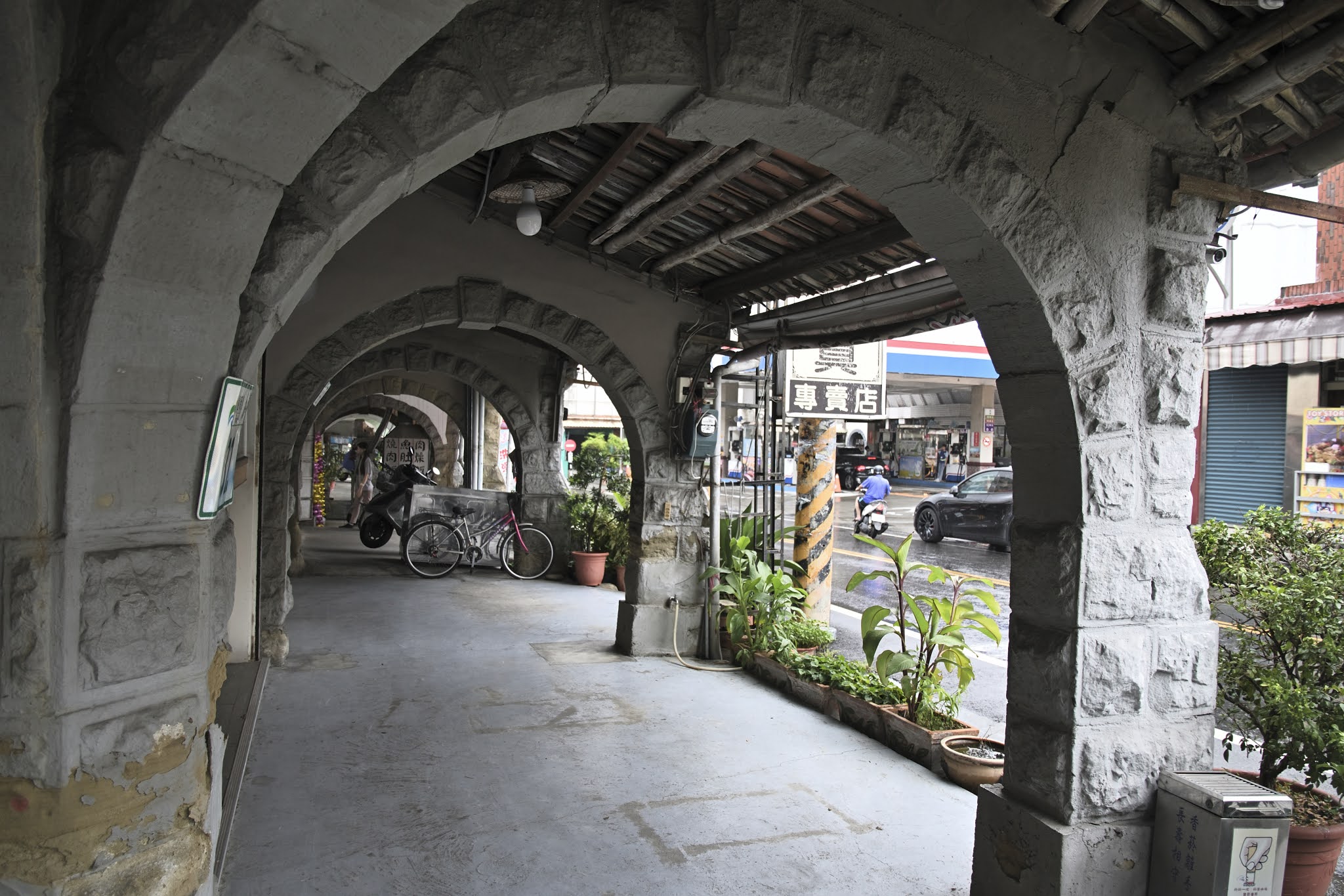 這張圖片展示一排具有拱型石造結構的騎樓,騎樓下方有單車、機車和許多盆栽,外頭則是車輛與商店街景。
