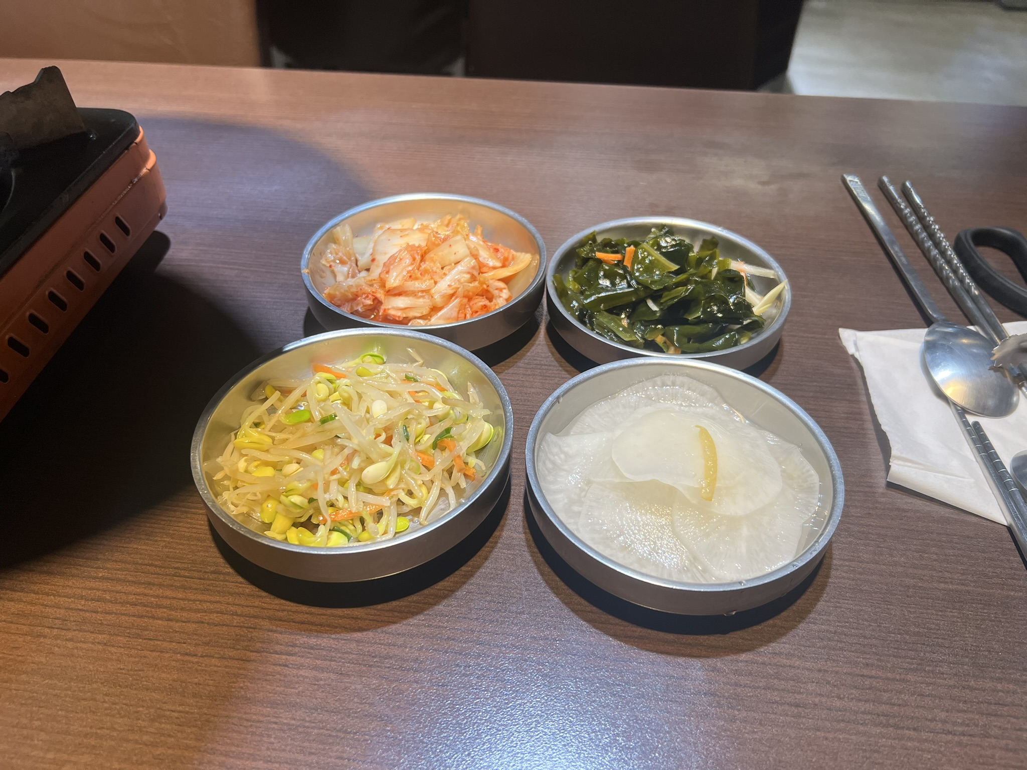 這張圖片主要有四款韓式小菜,分別是泡菜、涼拌海帶、涼拌豆芽和醃蘿蔔,擺放在圓形小盤中,桌面乾淨整齊,圖片中無明顯浮水印。