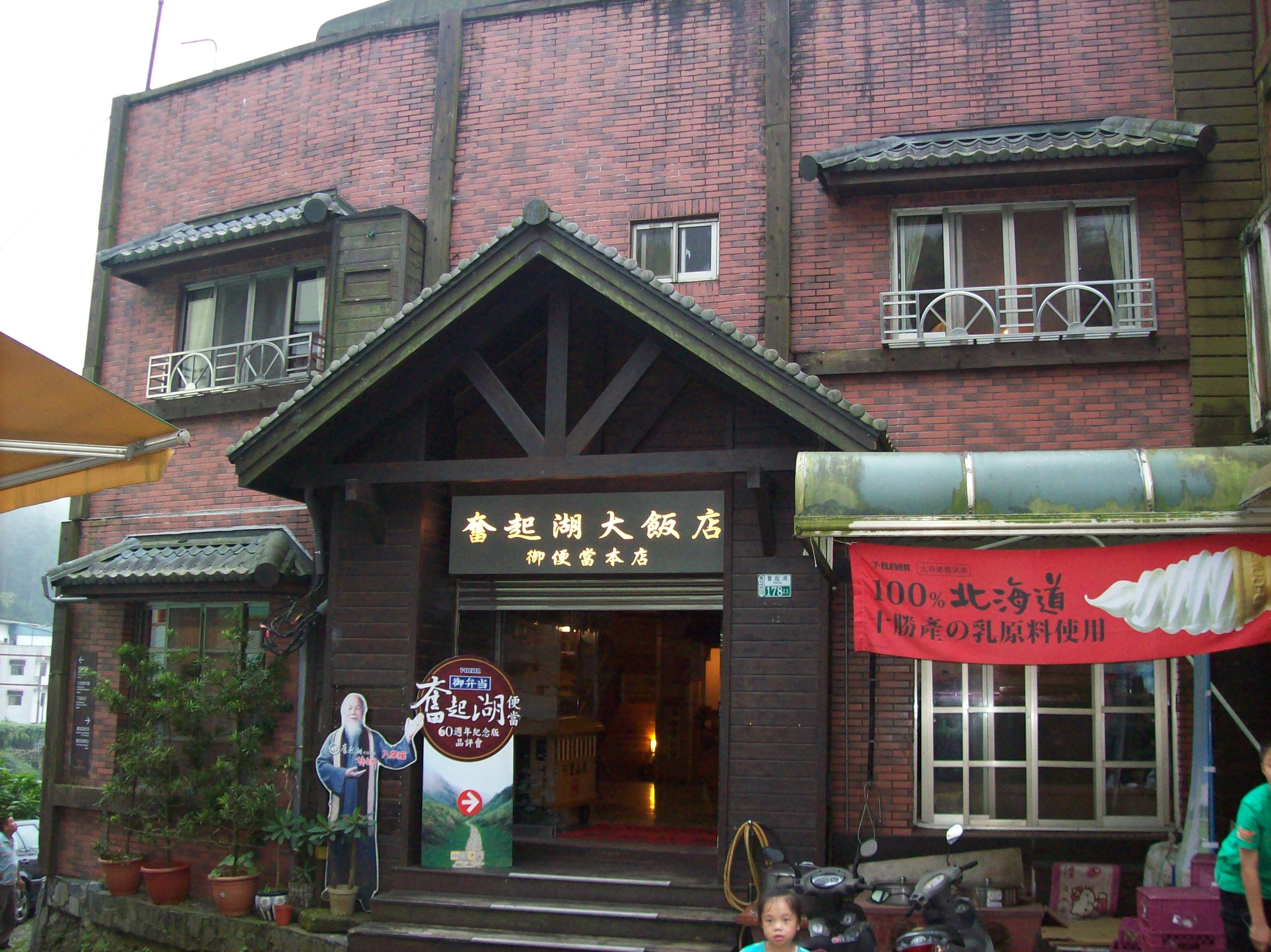 這張圖片是一家名為『春把湖大飯店』的建築正門,外牆為紅磚設計,入口上方有金色招牌,旁邊有冰淇淋廣告及多個盆栽,建築整體有濃厚的台灣鄉村氛圍。圖片無明顯浮水印。
