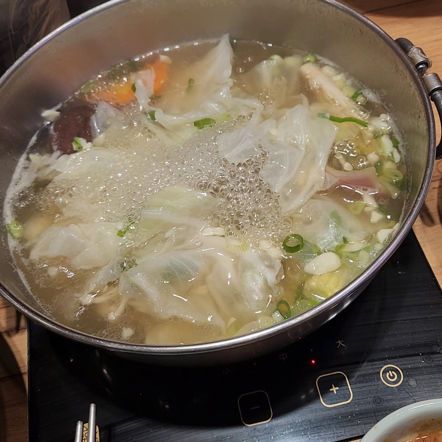 這張圖片是一鍋正在煮的火鍋,裡面有高麗菜、蔥花、香菇、紅蘿蔔等蔬菜,湯底正在加熱冒泡。圖片沒有浮水印。