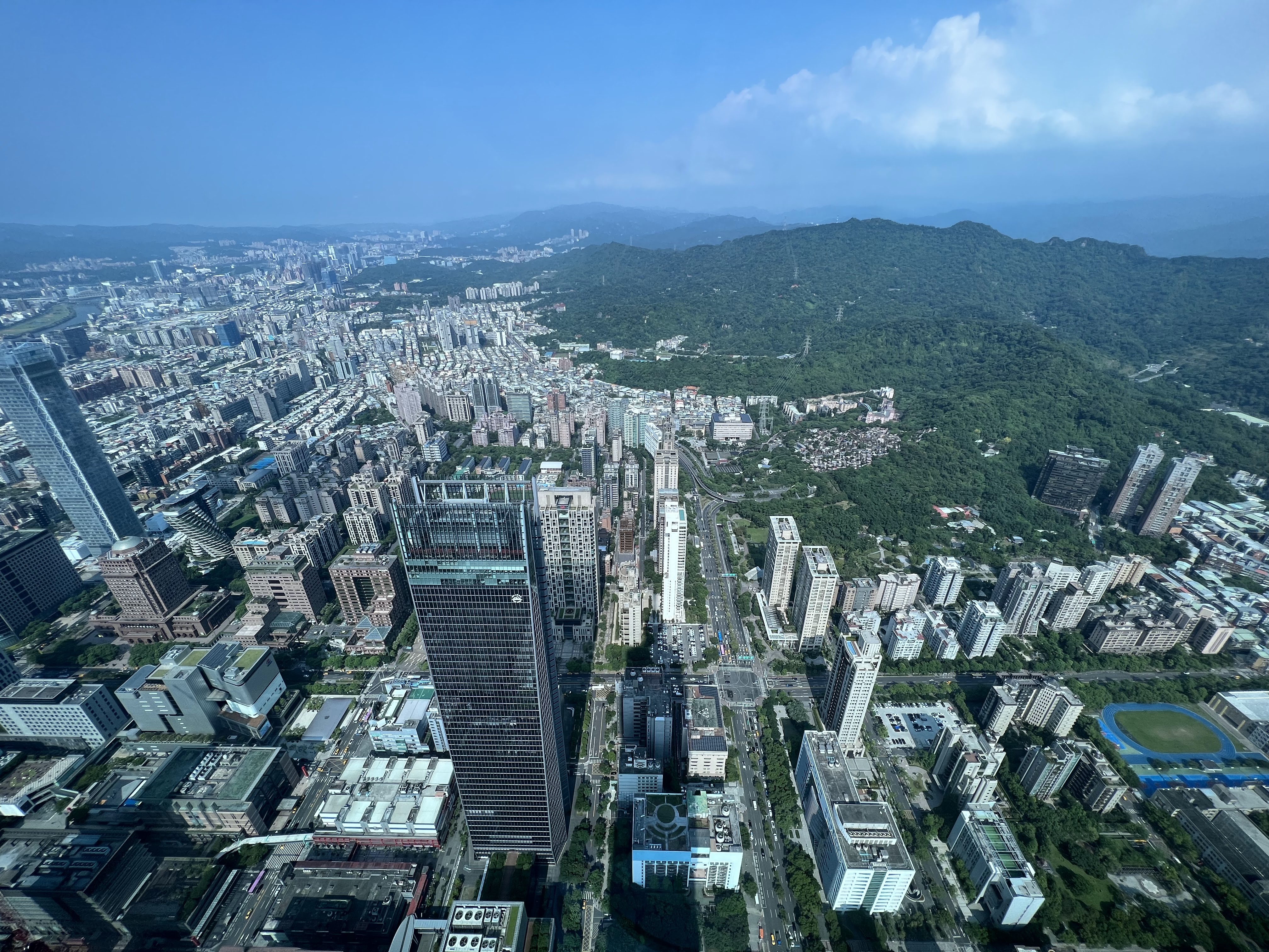 這張圖片展示一座城市的高空全景，畫面中有許多高樓大廈，遠方有大片綠色山林。天氣晴朗，藍天白雲，整體視野開闊，未見明顯浮水印。