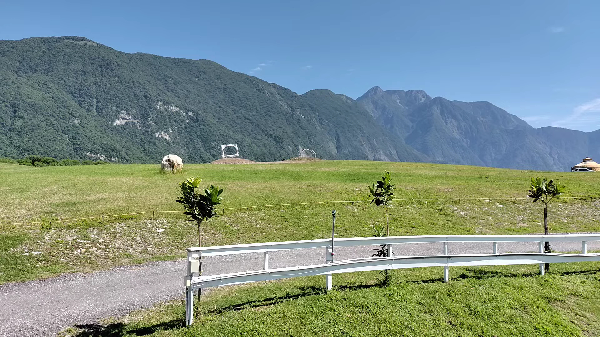廣闊綠色草原,遠處有高聳山脈和藍天,草原上有白色柵欄與裝置藝術