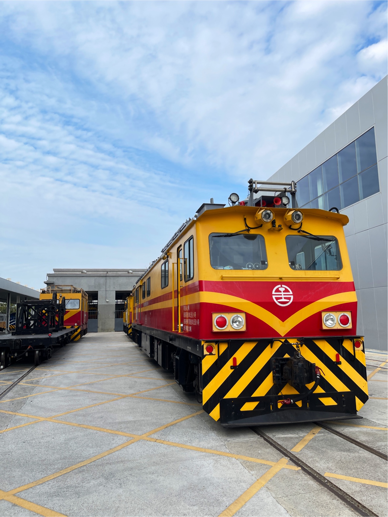 黃色和紅色相間的鐵道維修車停靠在戶外鐵軌上，背景有現代建築和藍天白雲。