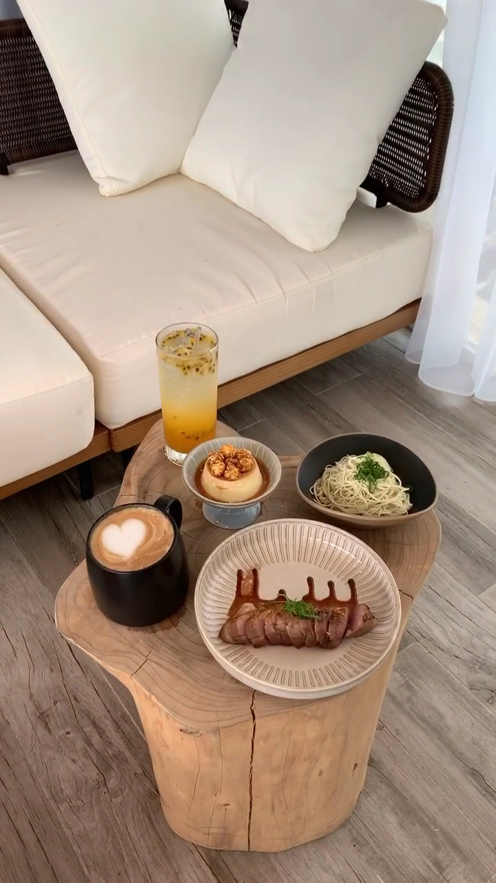 淺色沙發和原木茶几，桌上有拿鐵、冰飲、烤肉、義大利麵及布丁