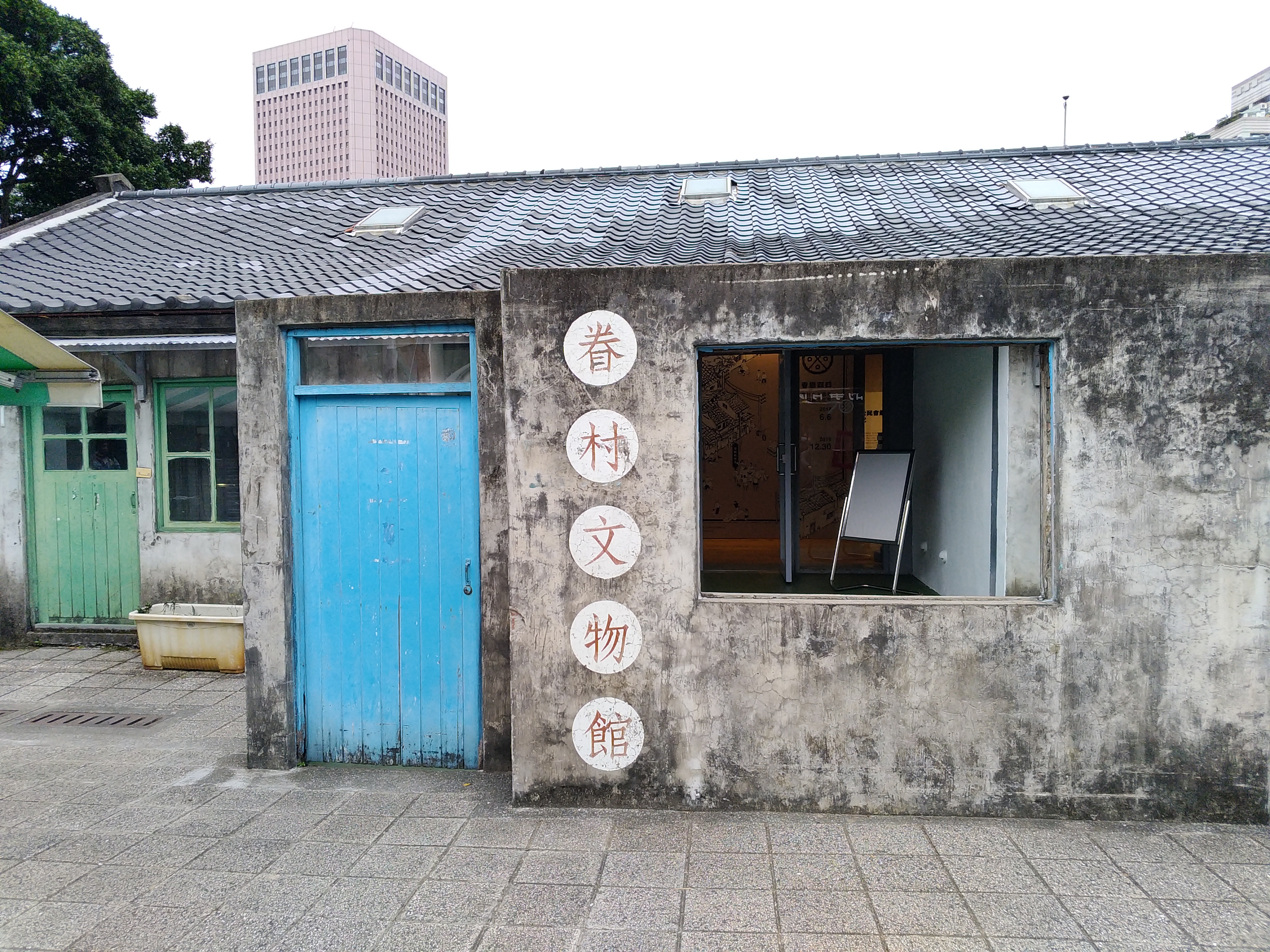 這張圖片主要是一棟老舊建築，牆上有『眷村文物館』的字樣。左側有藍色和綠色的門窗，建築外觀斑駁，中間有開放式的窗口可見室內展示空間，背景有高樓。圖片無明顯浮水印。