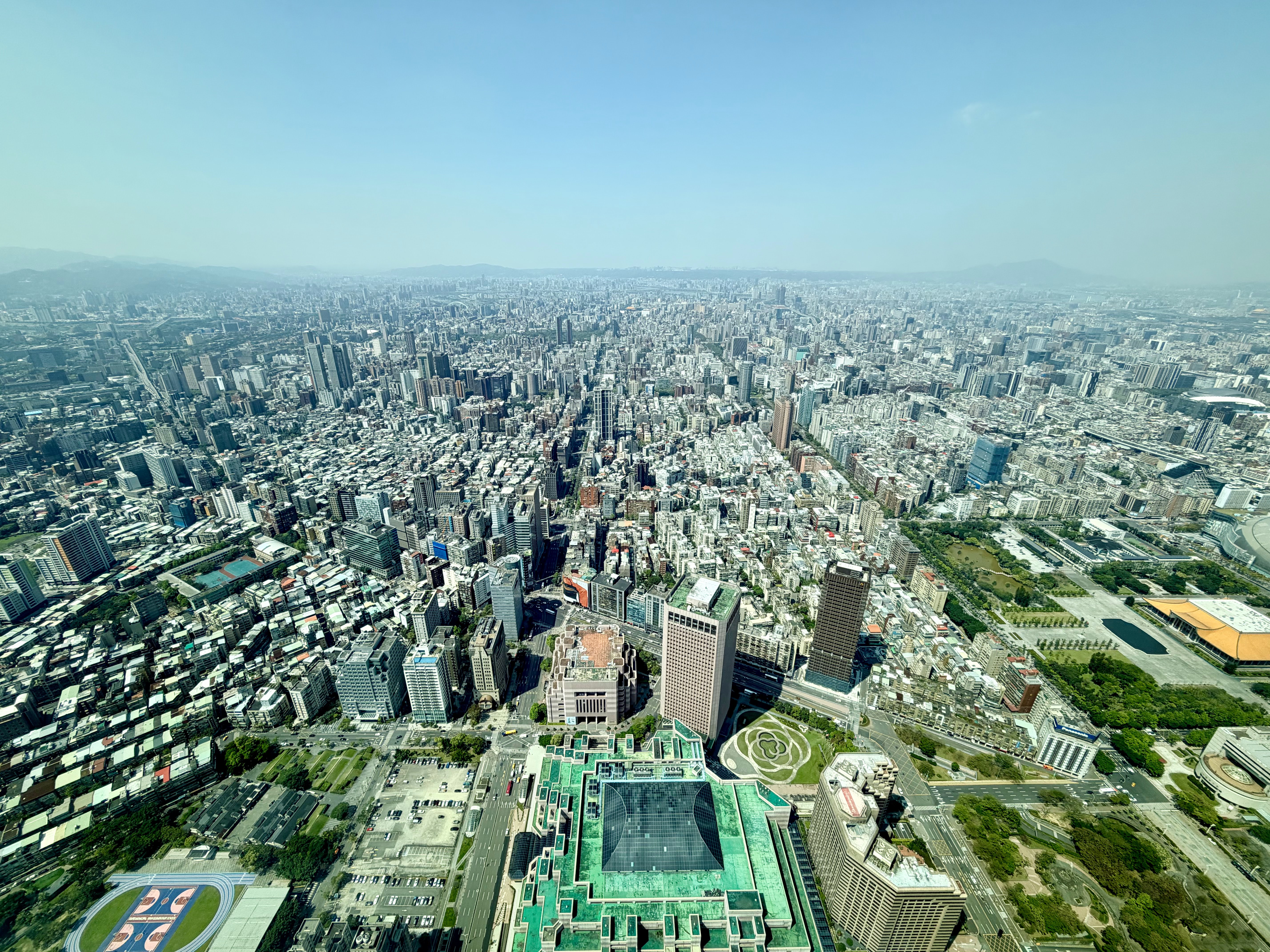 這張圖片是一座城市的高空俯瞰圖，畫面中可見密集的高樓大廈、馬路及綠地。遠方有山脈，天空晴朗，整體景觀繁華壯觀。圖片右下方還有明顯的體育場和樹林。圖片無浮水印。