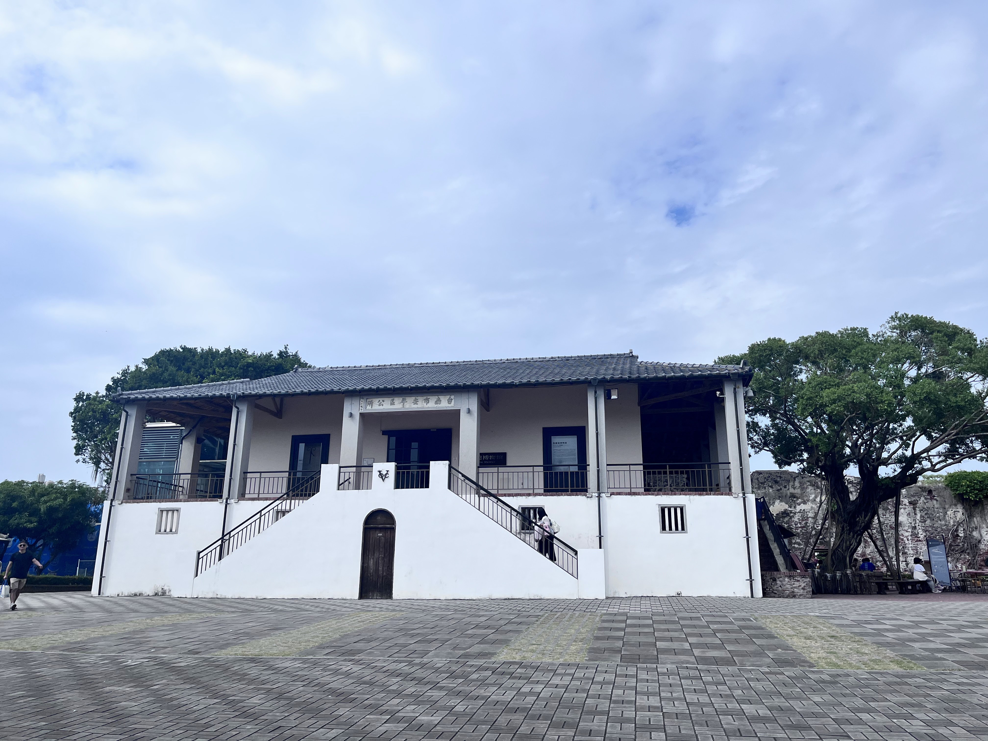 這張圖片主要是一棟兩層樓的白色建築,前方有大面積的廣場,背景有樹木與藍天,環境寬敞清新。圖片中未見明顯浮水印。