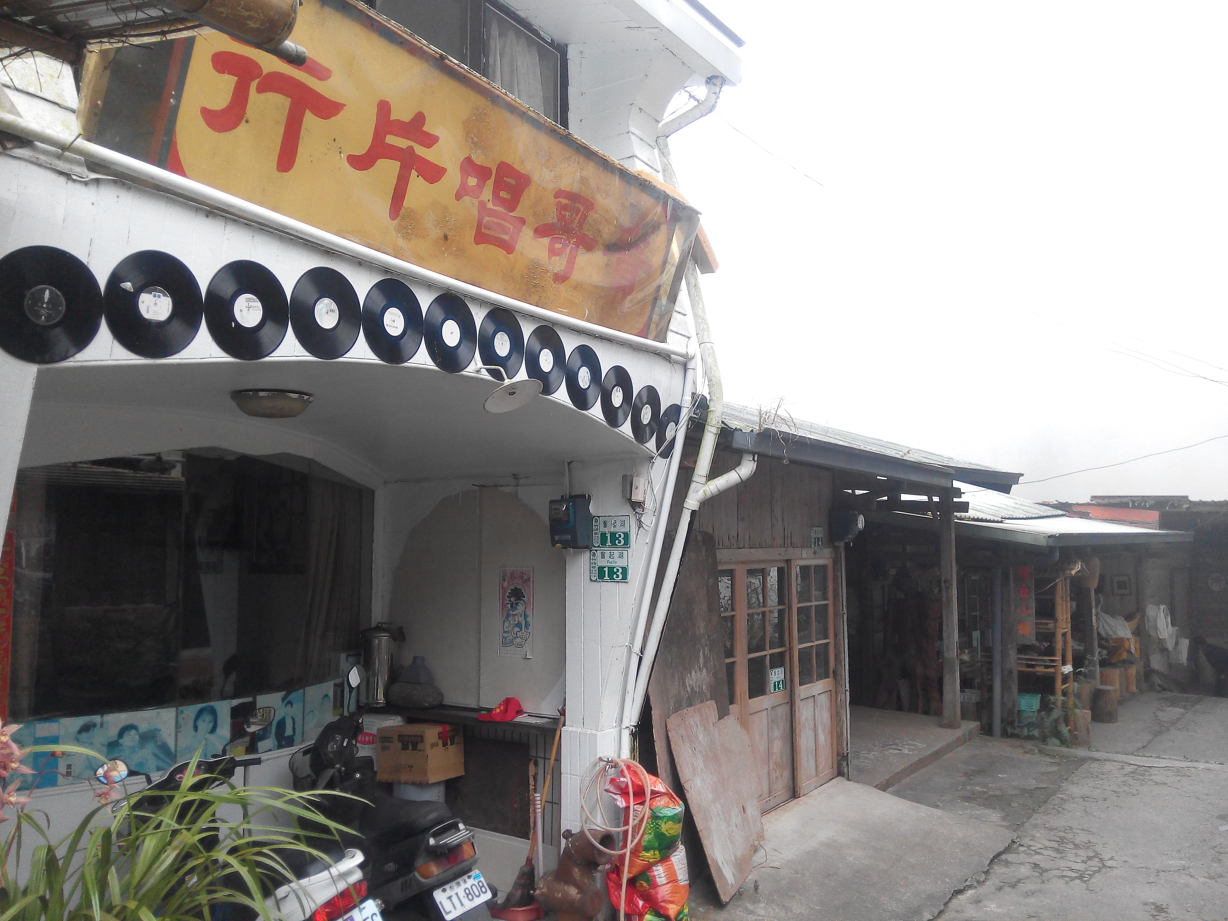 這是一家名為『斤片唱歌』的懷舊風格店家,建築外牆裝飾了許多黑膠唱片,門口有機車與植栽,旁邊有舊式木屋,未見明顯浮水印。