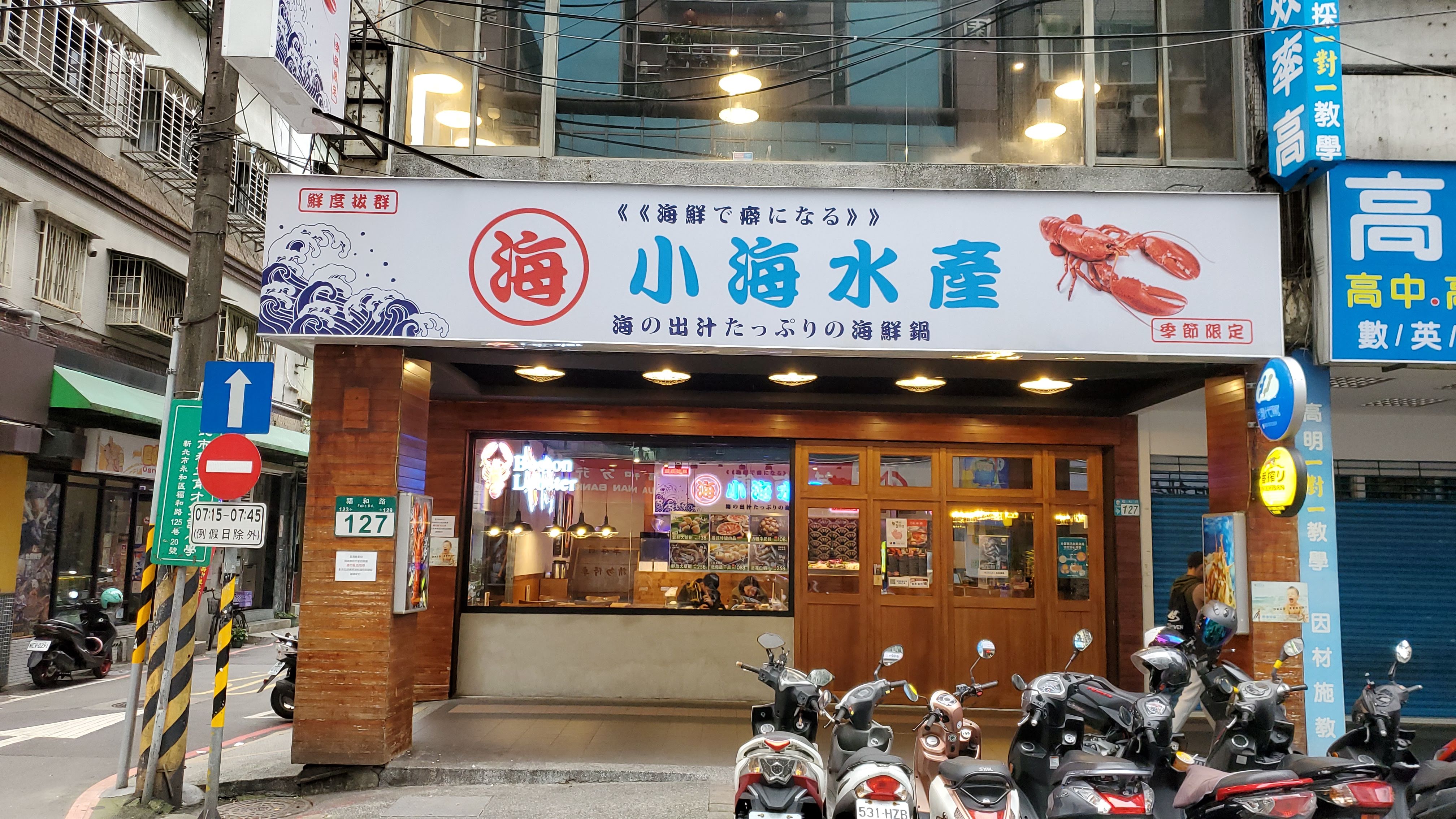 這張圖片主要是一家名為『小海水產』的海鮮餐廳門口,招牌上有大龍蝦圖案和日文標語,店外有機車停放,並沒有明顯浮水印。