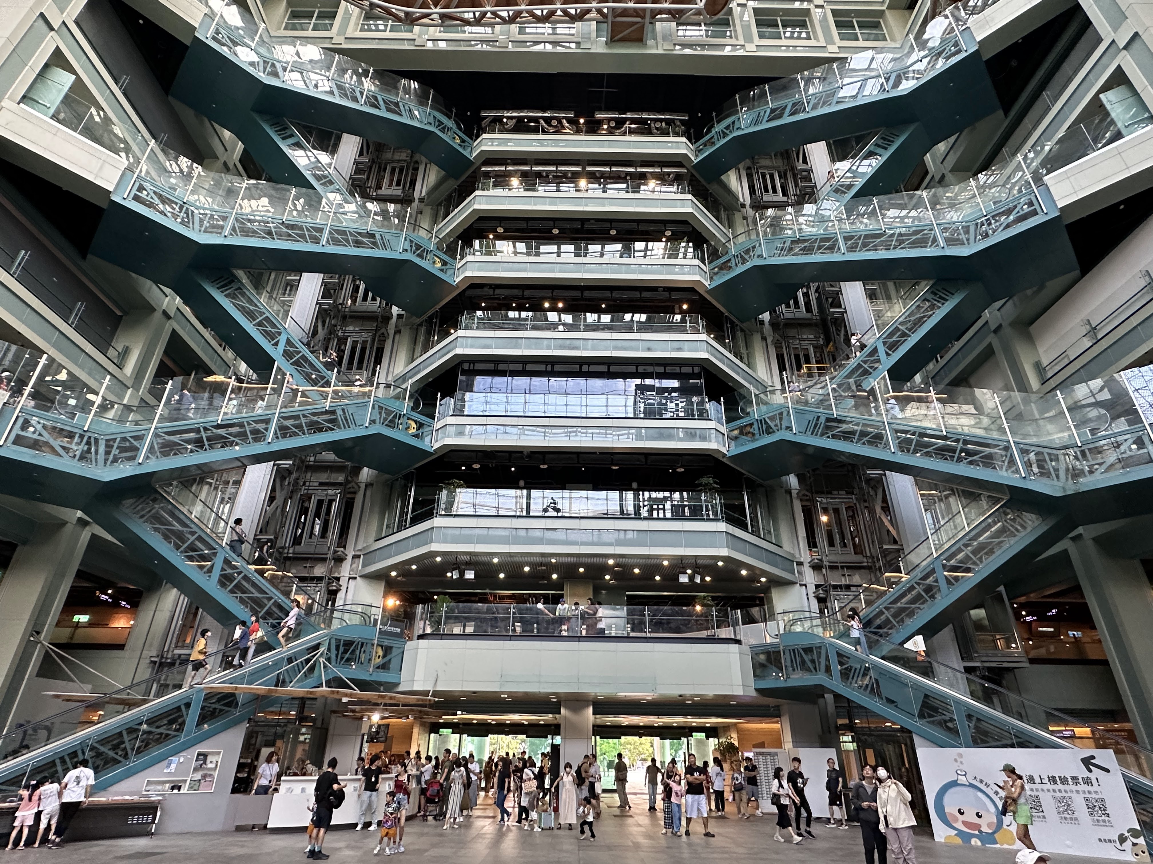 這張圖片展示了一座現代化的大型室內建築，中央有四層交錯延伸的藍綠色樓梯，許多民眾在樓梯與大廳移動。建築內部寬敞明亮，結構現代，沒有明顯浮水印。