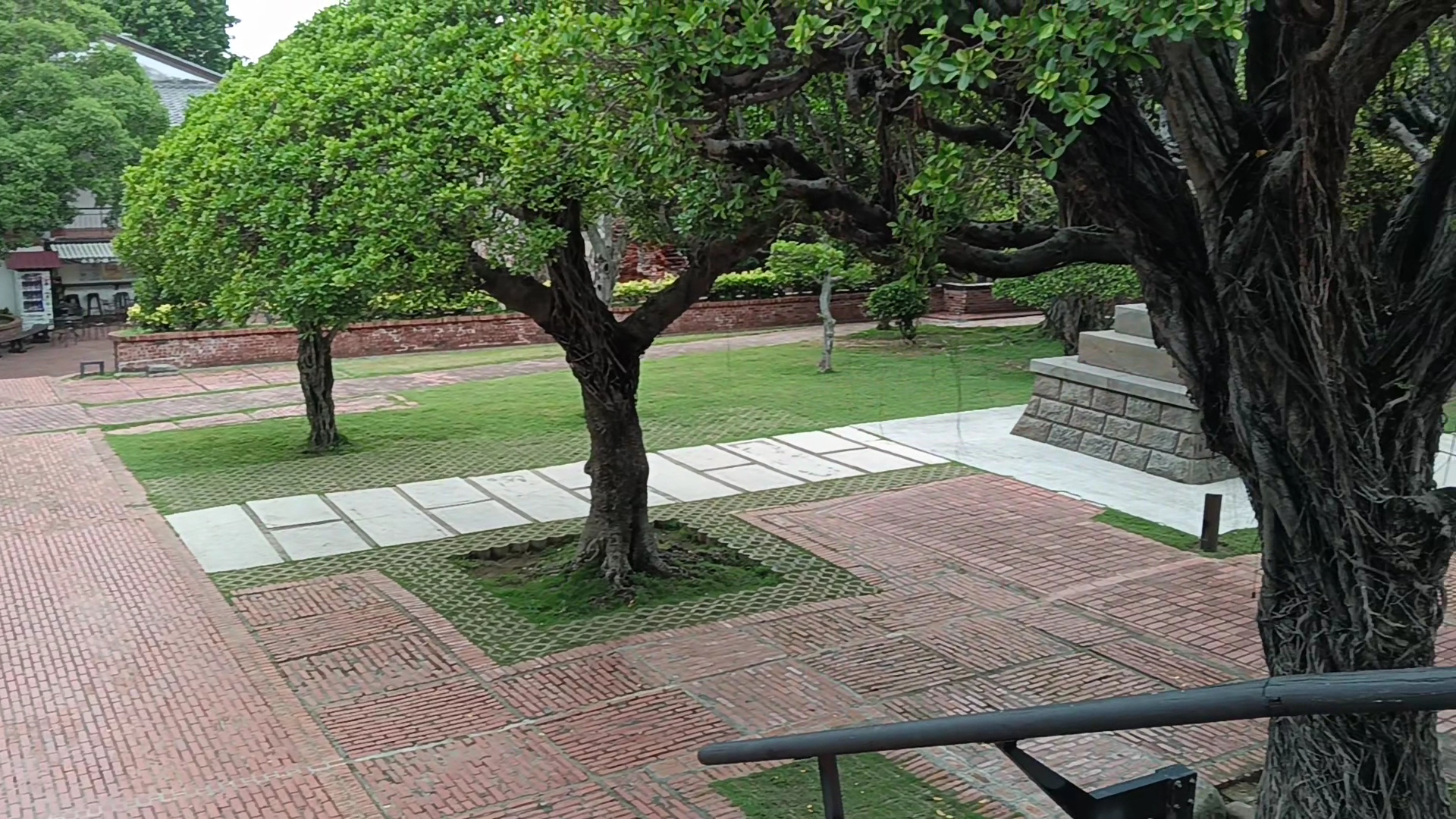 這張圖片主要呈現一座設有磚紅色地磚與石板步道的庭園,庭園內有多棵大樹與綠色草地,周圍有矮磚牆,並無浮水印。
