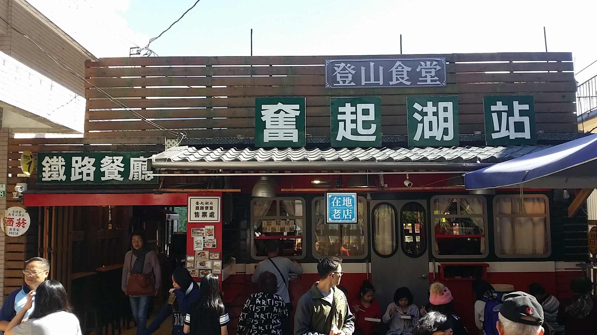 這張圖片主要呈現了台灣奮起湖車站旁的鐵路餐廳,有濃厚懷舊風格,外觀仿照火車車廂,門前有許多遊客聚集用餐及拍照,營造在地特色氣氛。圖片無明顯浮水印。