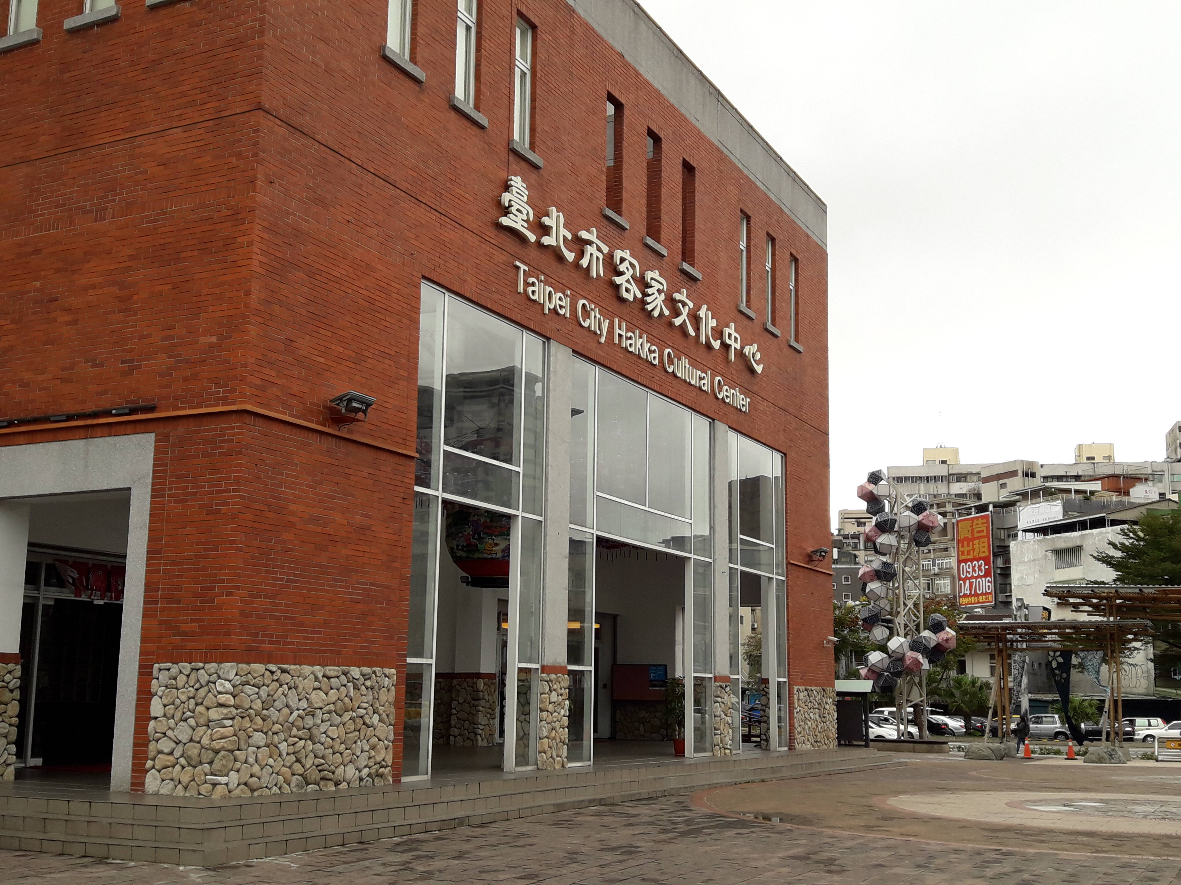 圖片主要顯示一棟紅磚建築，為台北市客家文化中心，其外牆下方有石頭裝飾，旁邊還有一個以傘組成的裝置藝術及一些公告招牌。圖片無明顯浮水印。
