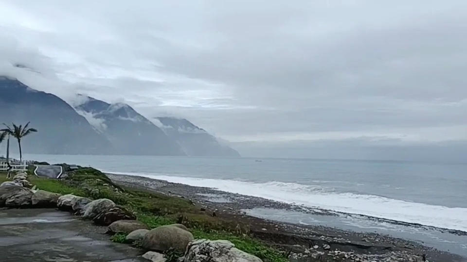 海邊風景,有海浪拍打岸邊,遠方有雲霧覆蓋的山脈與棕櫚樹