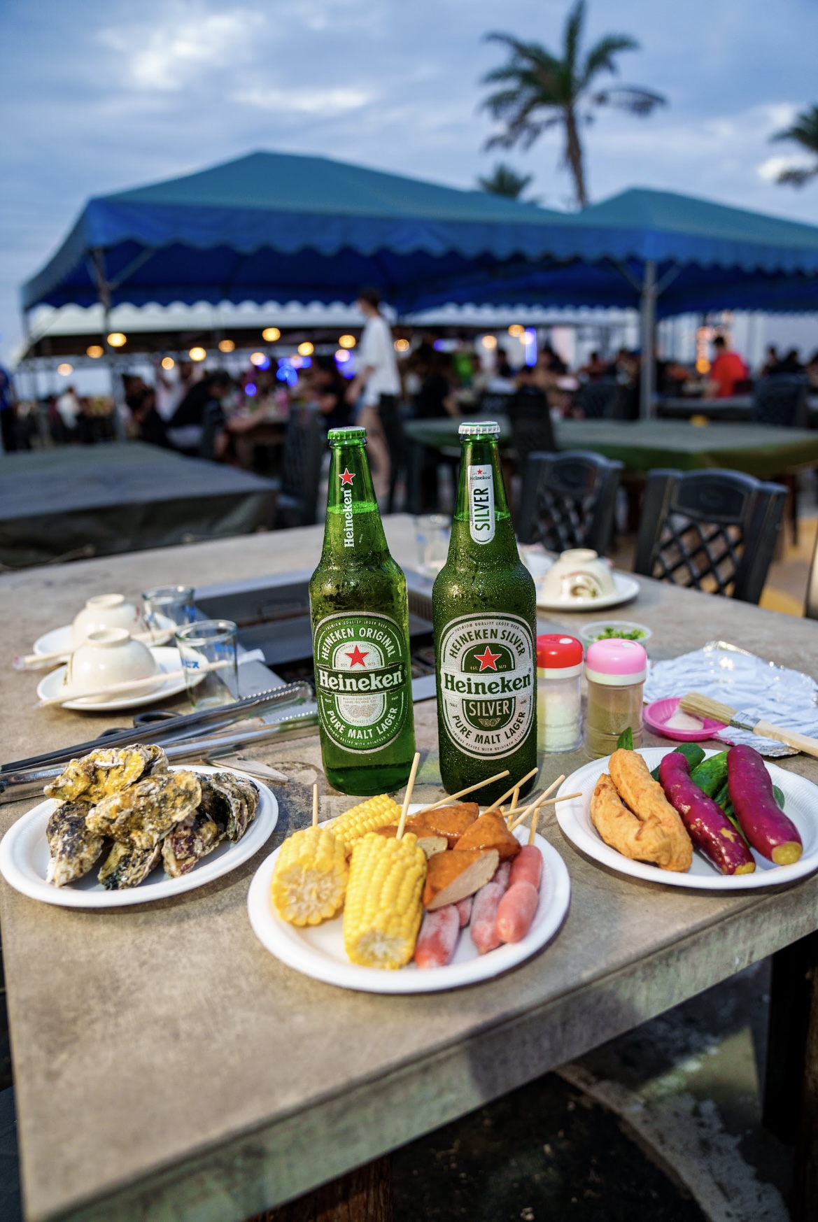 兩瓶海尼根啤酒與多盤燒烤食物，背景為戶外帳篷與棕櫚樹