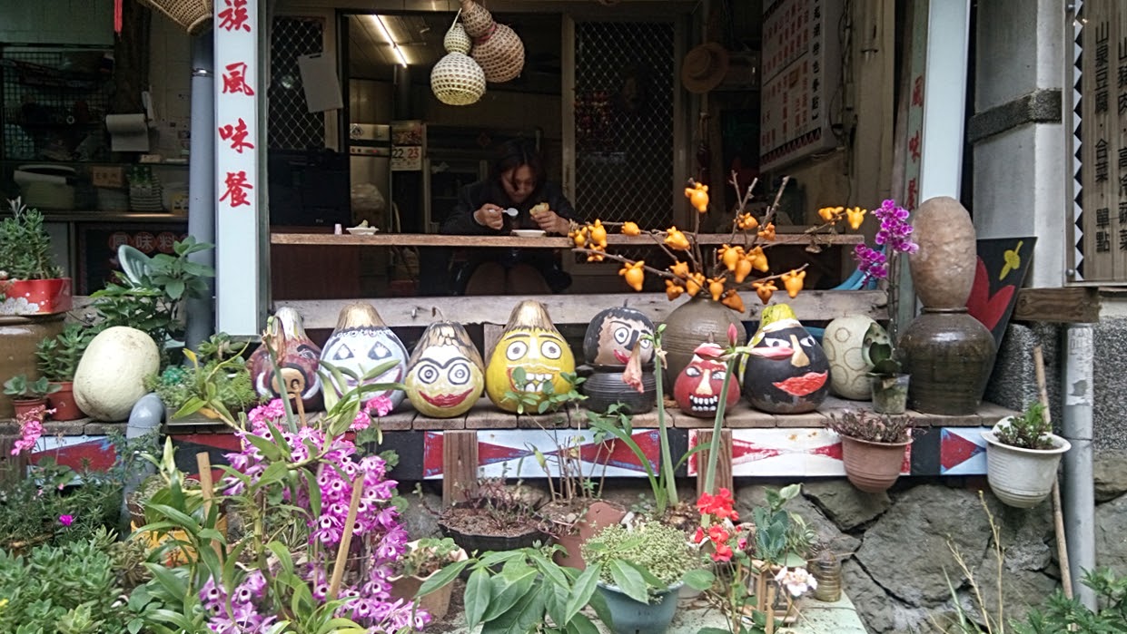 這張圖片主要展示了一家店的門口,有許多畫上表情的葫蘆和陶瓷裝飾,旁邊擺放著各種花卉與盆栽,整體氣氛充滿藝術感和生活氣息。畫面上無明顯浮水印。