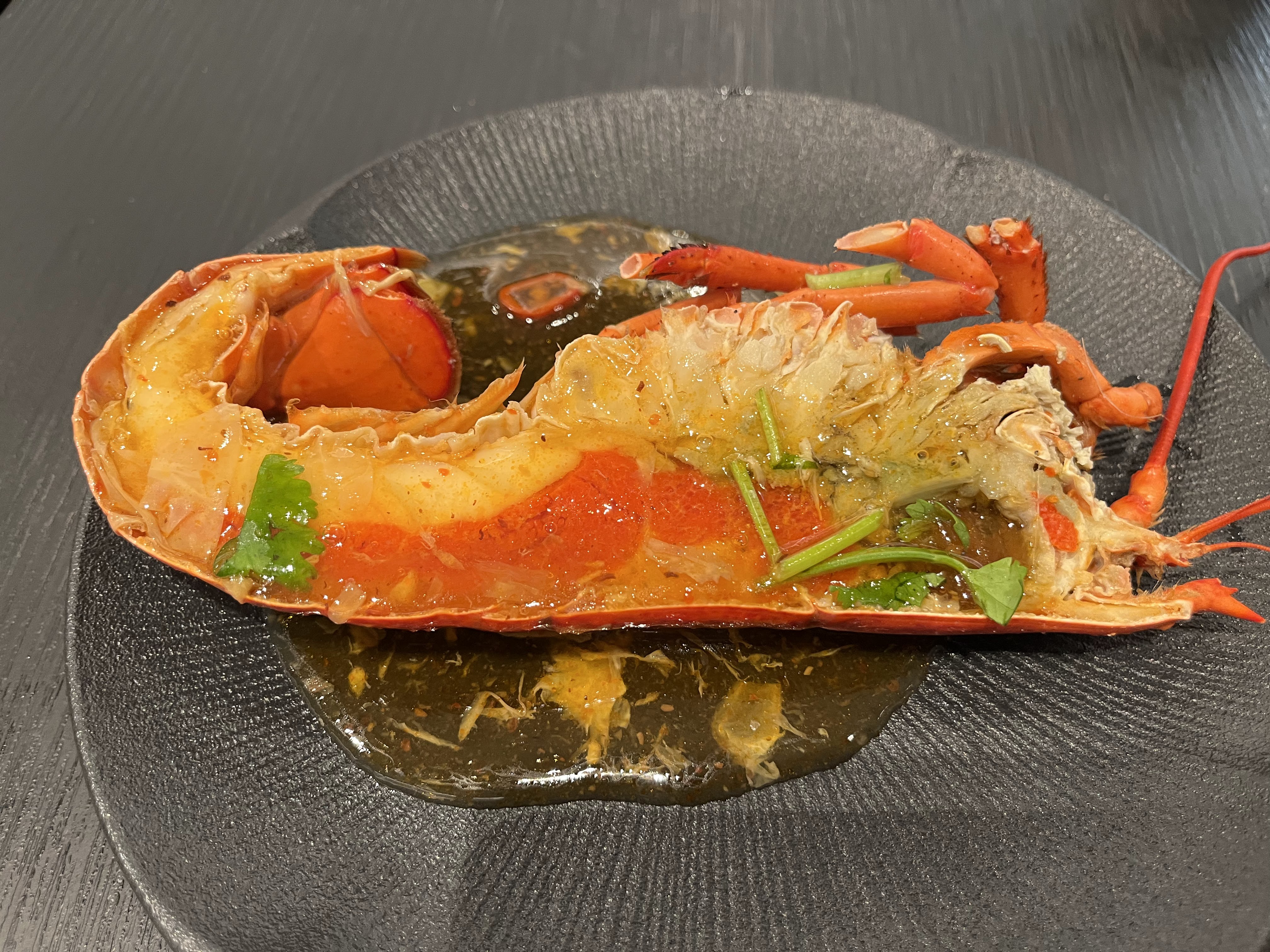 這張圖片展示了一道美食，一隻剖半的龍蝦，淋上醬汁並搭配香菜，擺放在黑色盤子上，呈現新鮮且誘人的樣貌。圖片無浮水印。