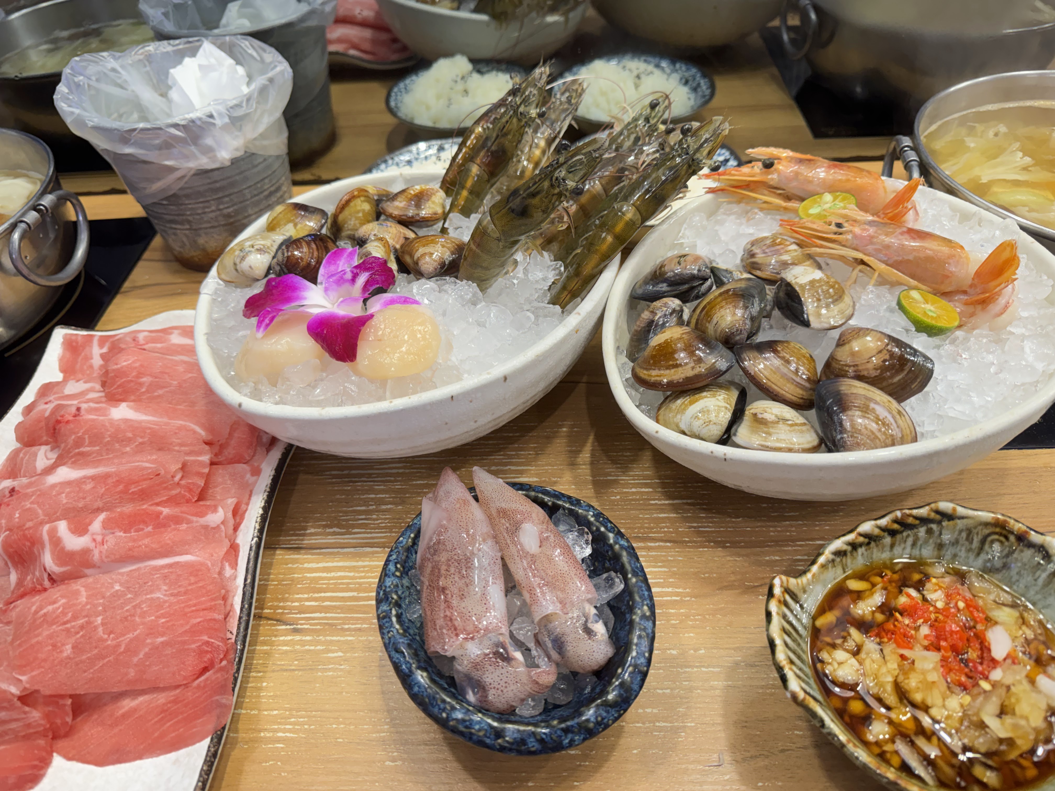 這張圖片展示了一桌新鮮火鍋食材,包括牛肉片、蛤蜊、鮮蝦、干貝、小章魚,以及沾醬和其他配料。海鮮擺在碎冰上,保持新鮮感。未見浮水印。