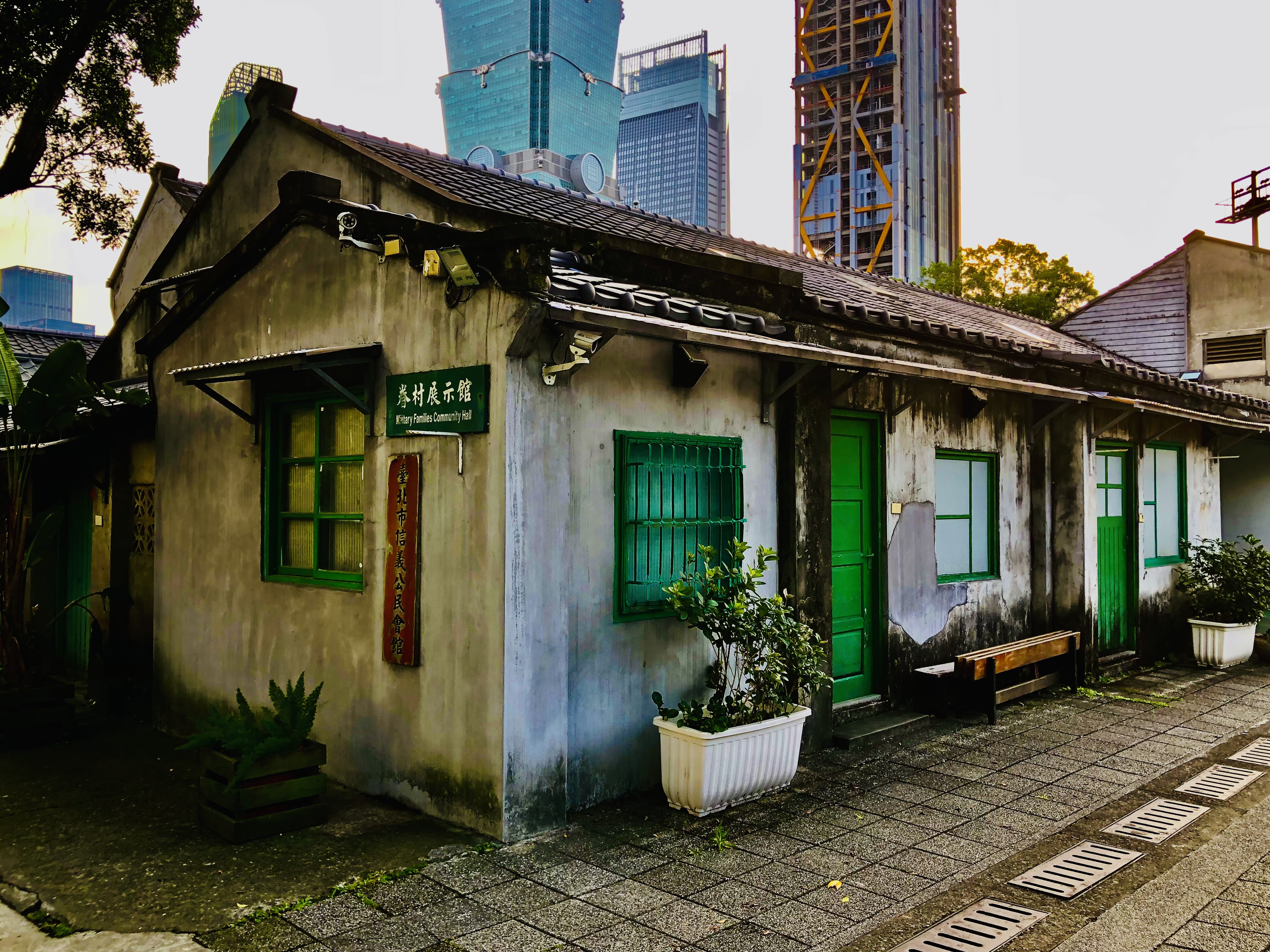這張圖片主要是一棟老式眷村建築，正面有綠色窗戶和大門，旁邊有盆栽，牆上掛有『眷村居民示範館』的招牌。背景則有現代高樓大廈，呈現新舊建築的對比。圖片無浮水印。