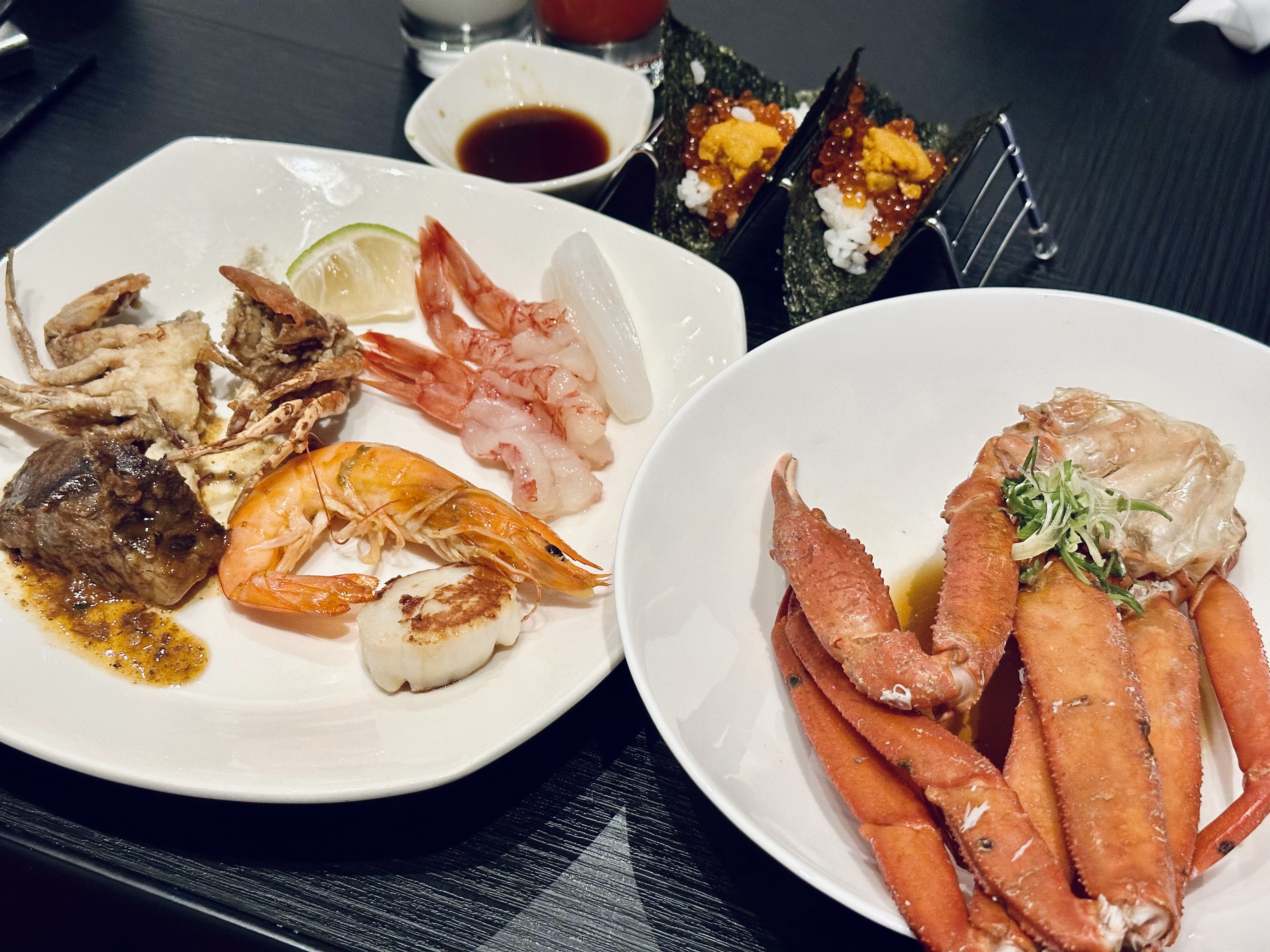 這張圖片主要有各式海鮮料理，例如蟹腳、蝦子、干貝和生魚片，還有手卷及牛肉。食物擺盤精美，沒有浮水印。