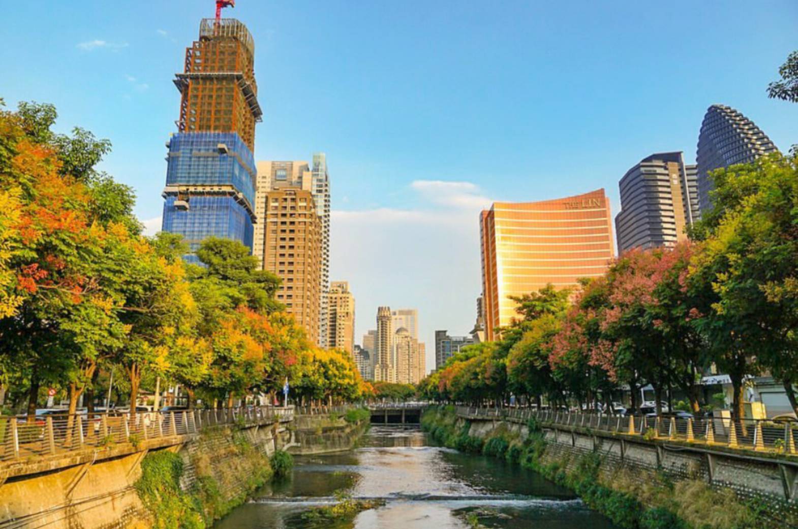 這張圖片展示一條河川穿過城市,兩旁有綠樹與秋色楓葉,遠方有許多現代高樓和正在興建的大樓。建築物間的藍天讓整體明亮宜人。圖片無明顯浮水印。