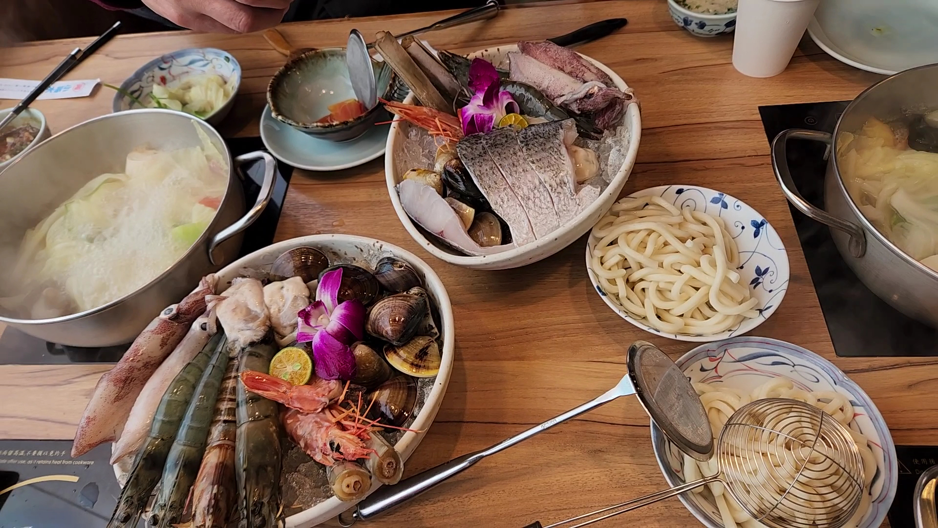 這張圖片主要展示了豐盛的火鍋料理,包括多種新鮮海鮮如蝦、魚、花枝、貝類並有烏龍麵和蔬菜。畫面色彩豐富,有美麗的紫色花朵裝飾,食材擺盤精美,看起來十分美味。圖片中無明顯浮水印。