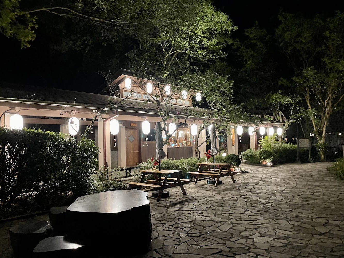 夜晚戶外餐廳掛滿日式燈籠,前方有木製桌椅與石椅