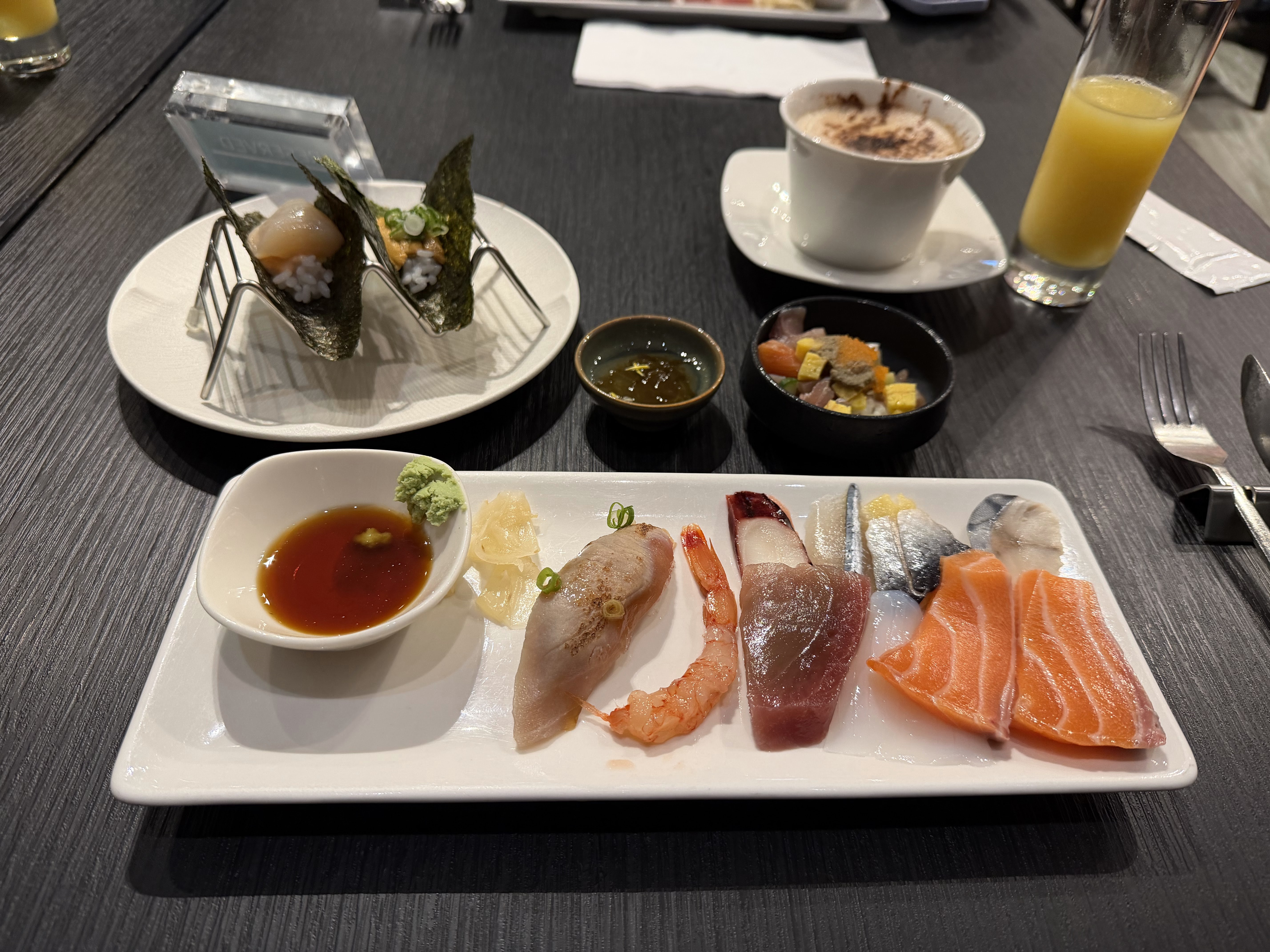 圖片主要是日式料理，包括多款生魚片、手卷、以及小碟前菜、蘸醬和一杯果汁，還有一杯熱飲。桌面整潔，生魚片新鮮多樣，另有海苔手卷和甜點。無浮水印。