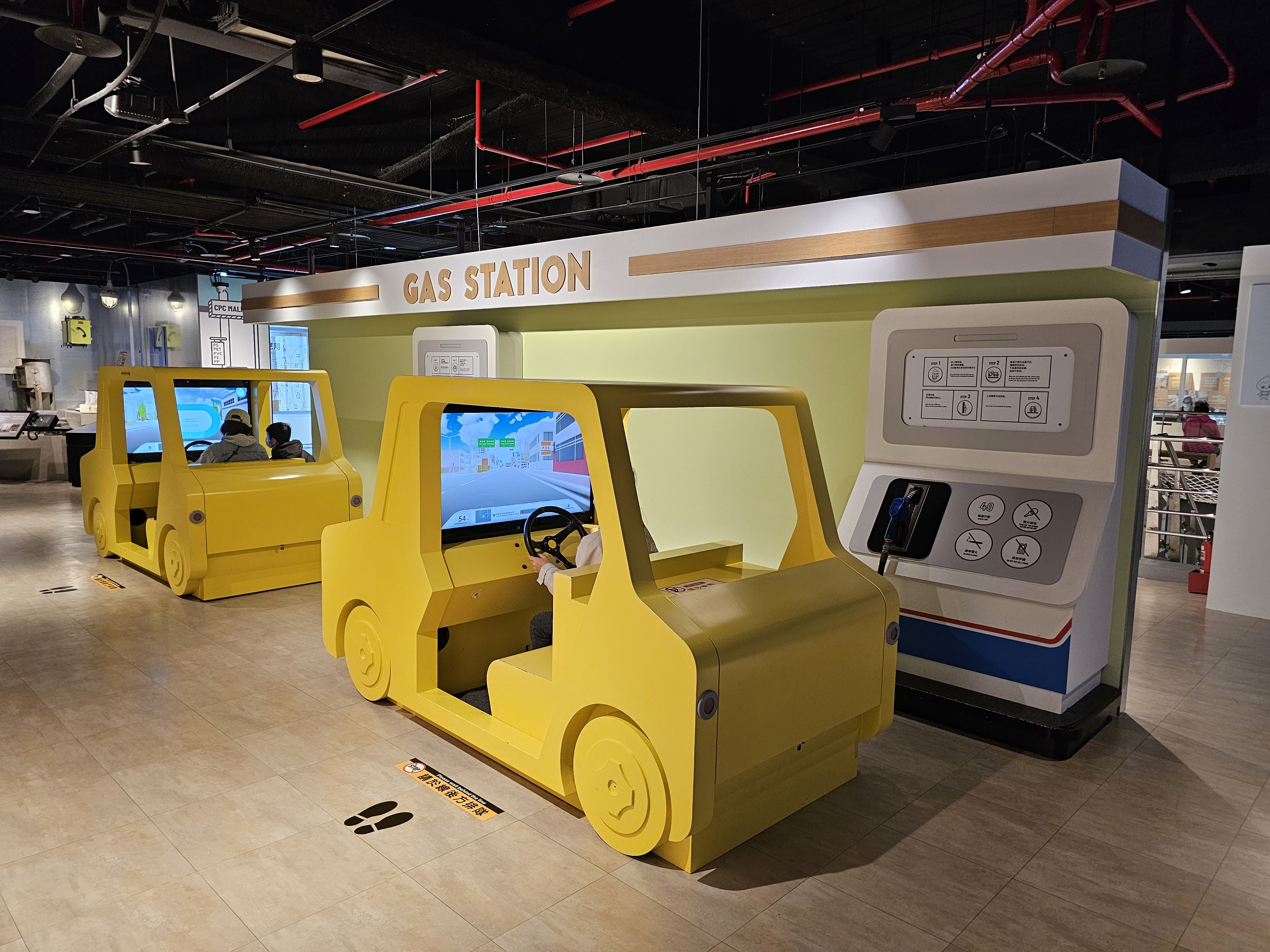 這張圖片拍攝於室內兒童遊樂區，有兩台黃色玩具車模擬駕駛器，前方模擬加油站設施。現場有小朋友坐在車內玩耍。圖片無浮水印。