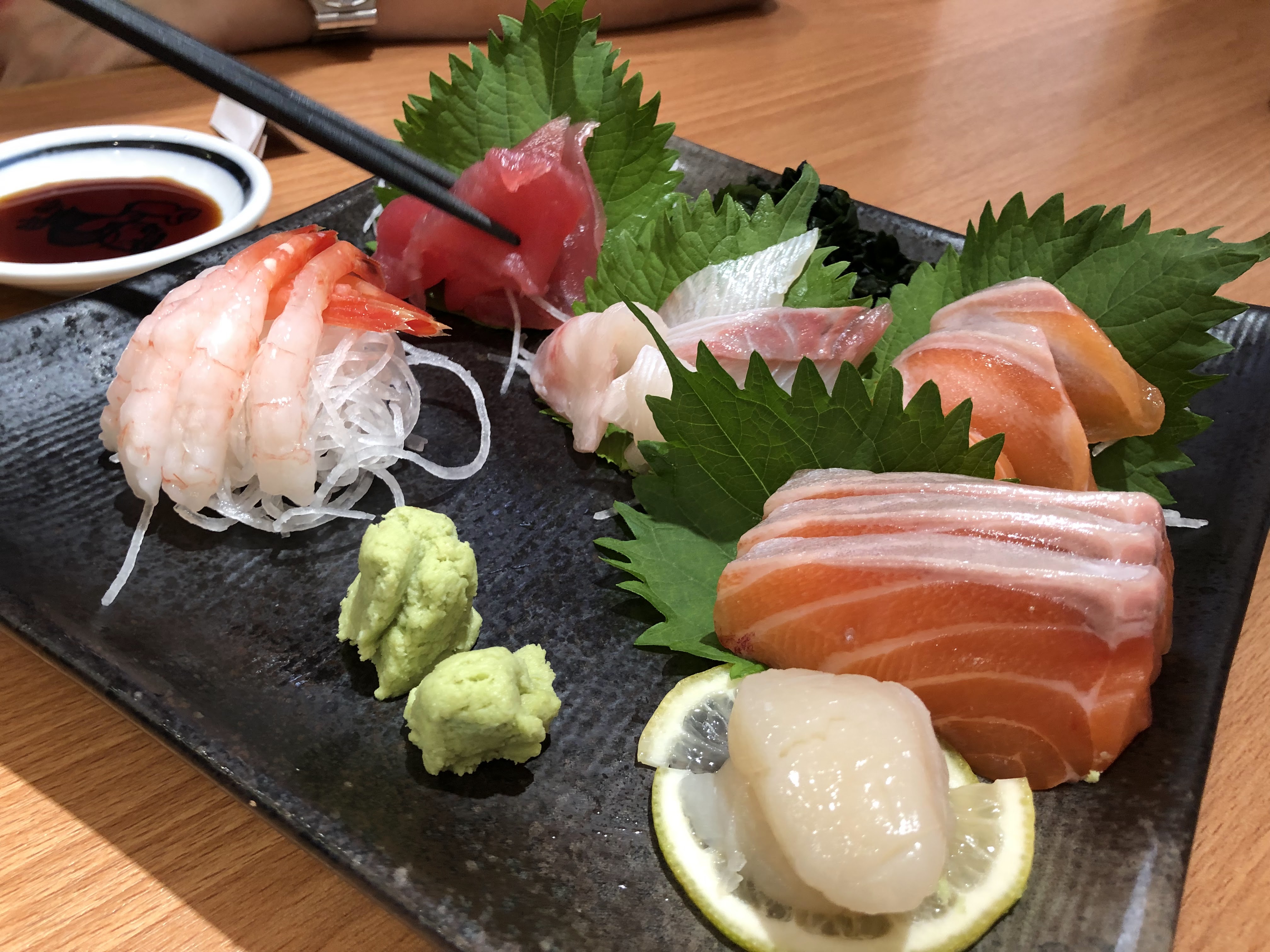 這張圖片主要是一盤日式刺身，包含鮭魚、鮪魚、甜蝦、干貝及白身魚切片，搭配蘿蔔絲、綠葉裝飾及芥末。旁邊有一小碟醬油，畫面中無明顯浮水印。