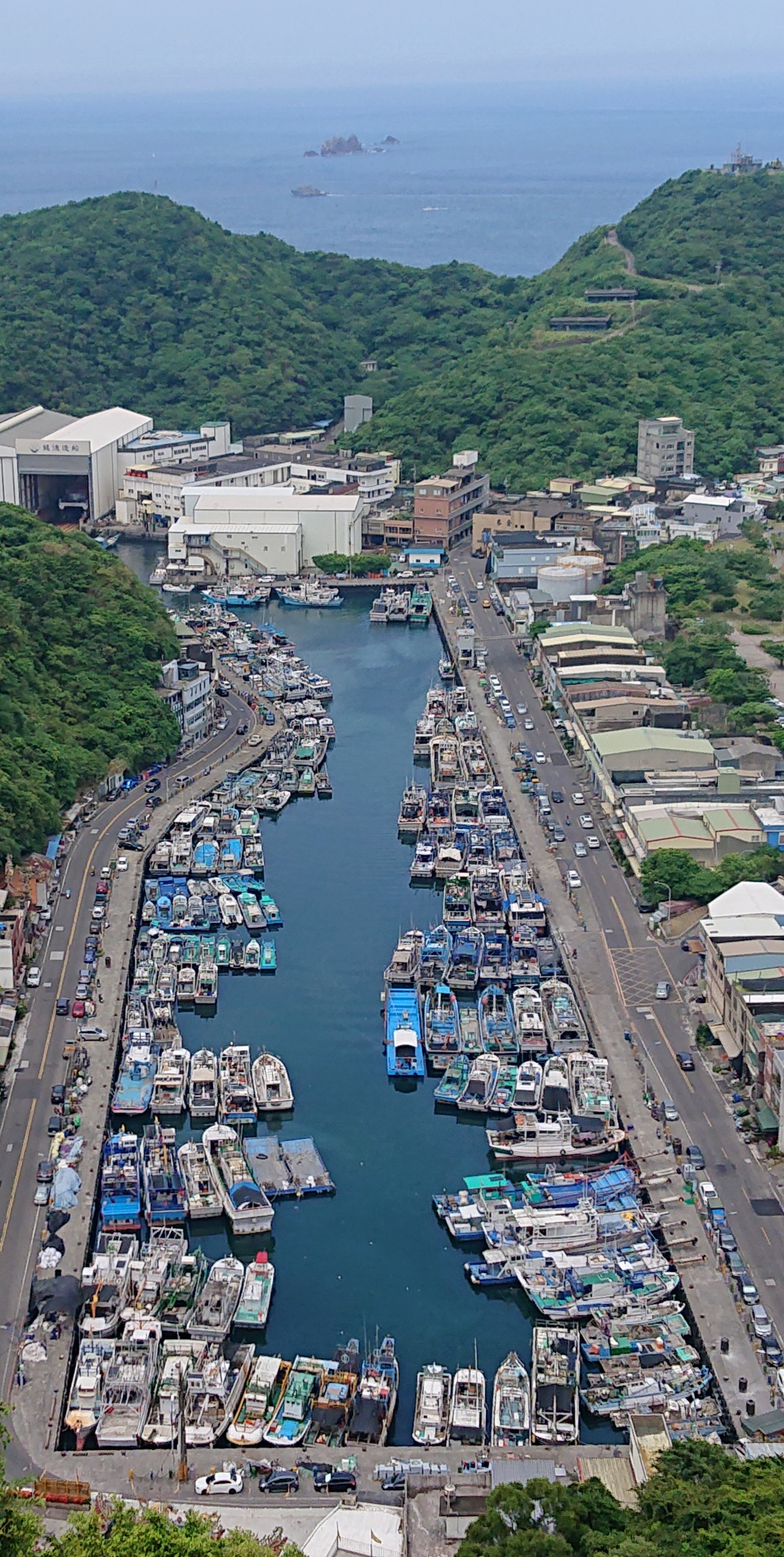 這張圖片主要是一個漁港，港內停滿了各式漁船，兩側為道路與建築物，周圍被青山環繞，遠方可以看到藍色海洋和幾座離岸岩石，圖中無明顯浮水印。