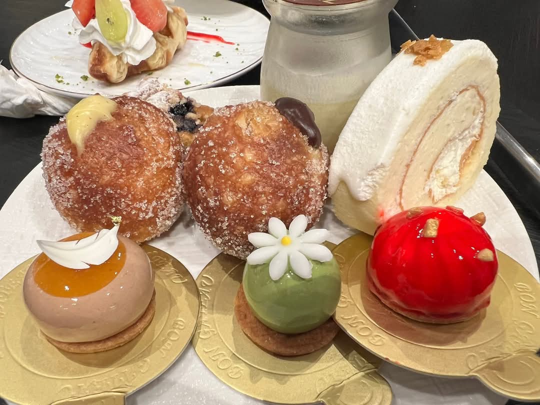 這張圖片展示了多款精緻甜點，包括泡芙、奶油卷、三種外觀繽紛的慕斯小蛋糕及一杯瓶裝甜品。甜點色彩豐富且擺盤精美，展現高級感。圖片右下角無明顯浮水印。