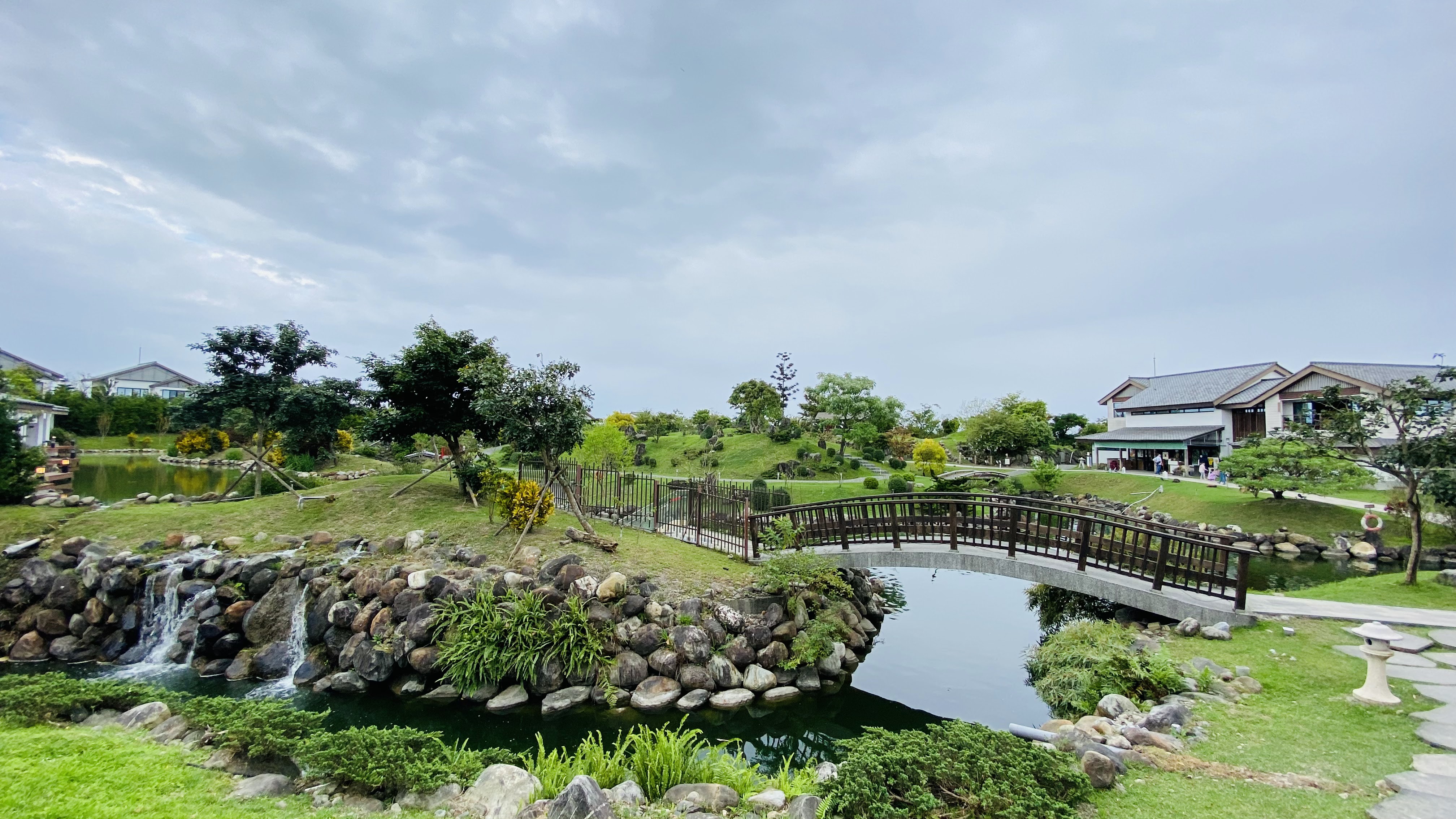 這張圖片主要是一個寬廣的綠意花園,中心有拱形橋橫跨池塘,水中有瀑布,四周種滿樹木與花草,右邊有建築物,營造出寧靜舒適的自然景色。圖片無明顯浮水印。